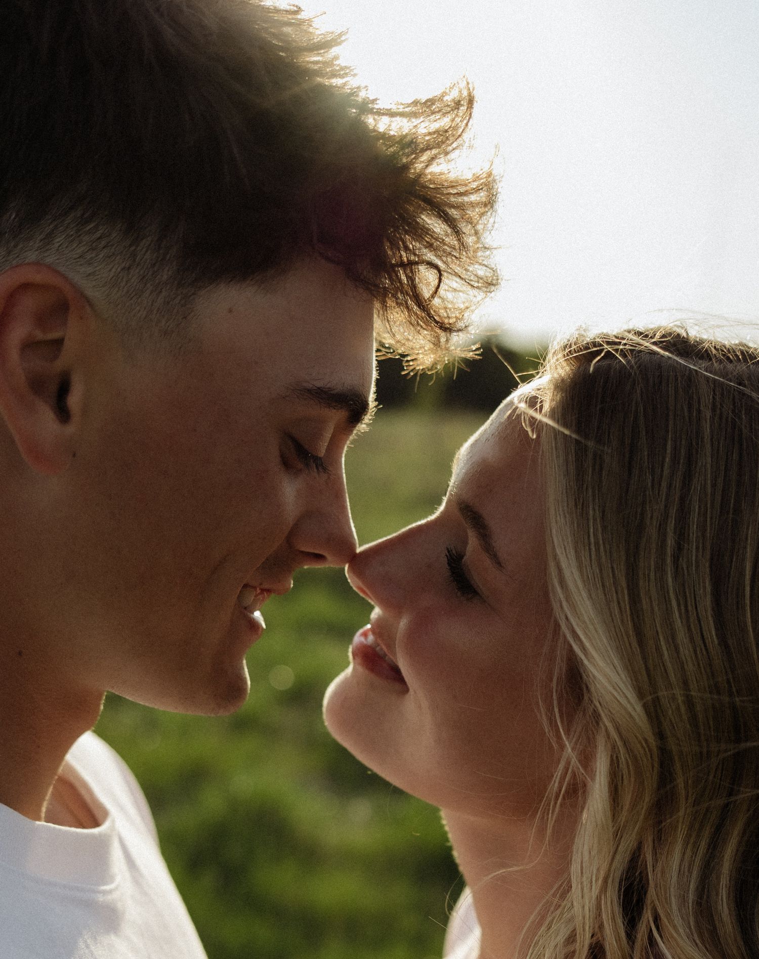 A young couple shares a romantic moment, noses almost touching, smiling softly in warm sunlight outdoors.