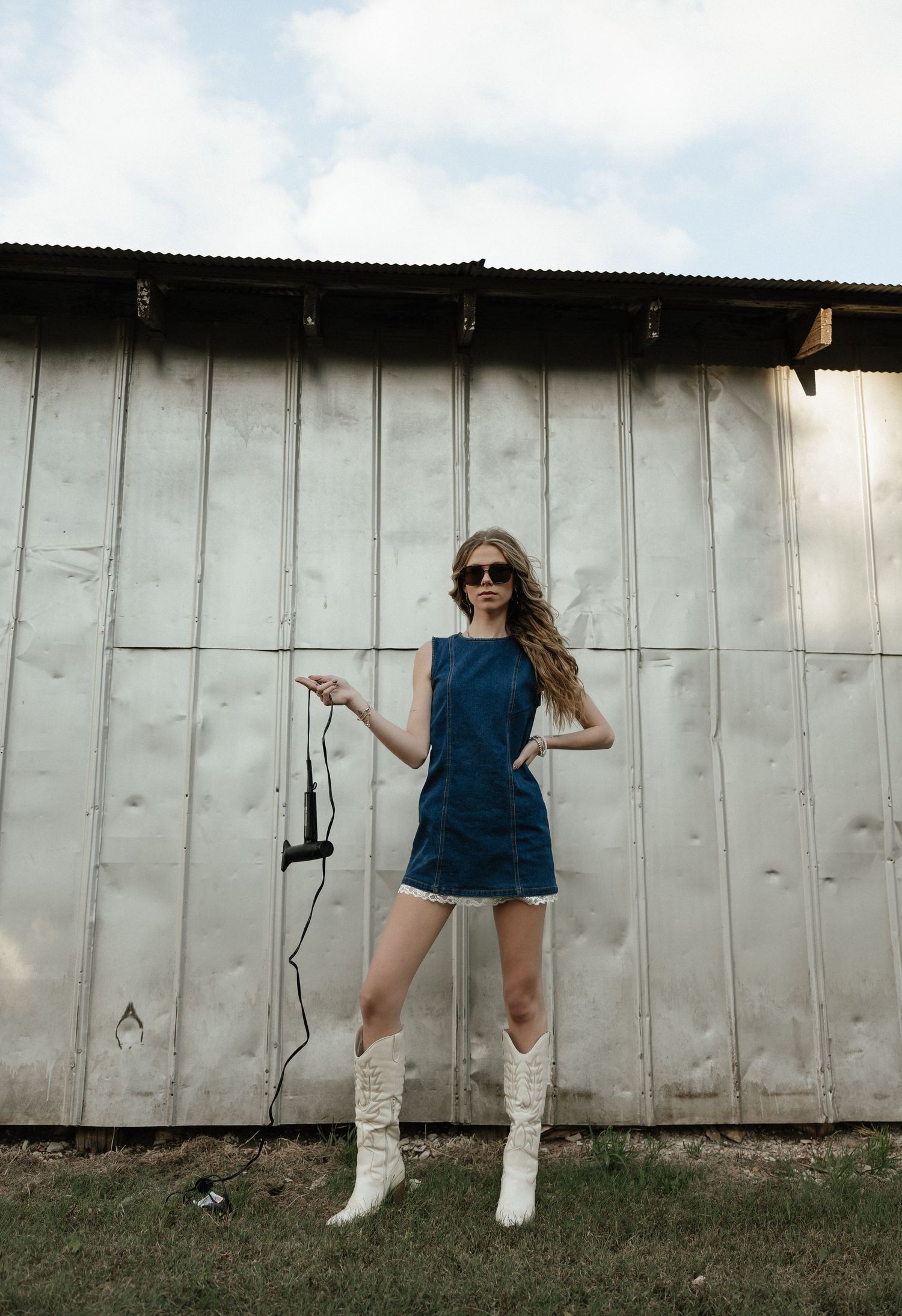 Woman in denim mini dress, white cowboy boots, and sunglasses poses outdoors holding a camera strap by a metal barn wall.