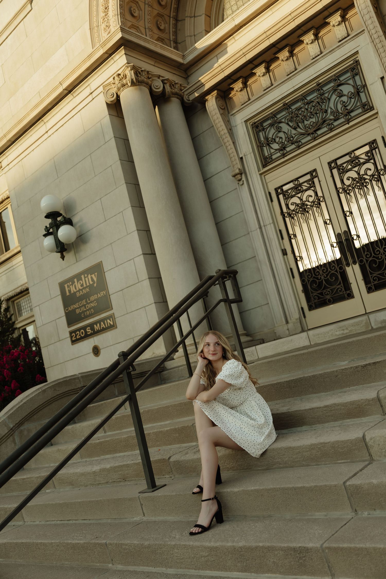 Young woman in white dress sitting on steps of classic Fidelity building at 220 E Main, posing for portrait photo.