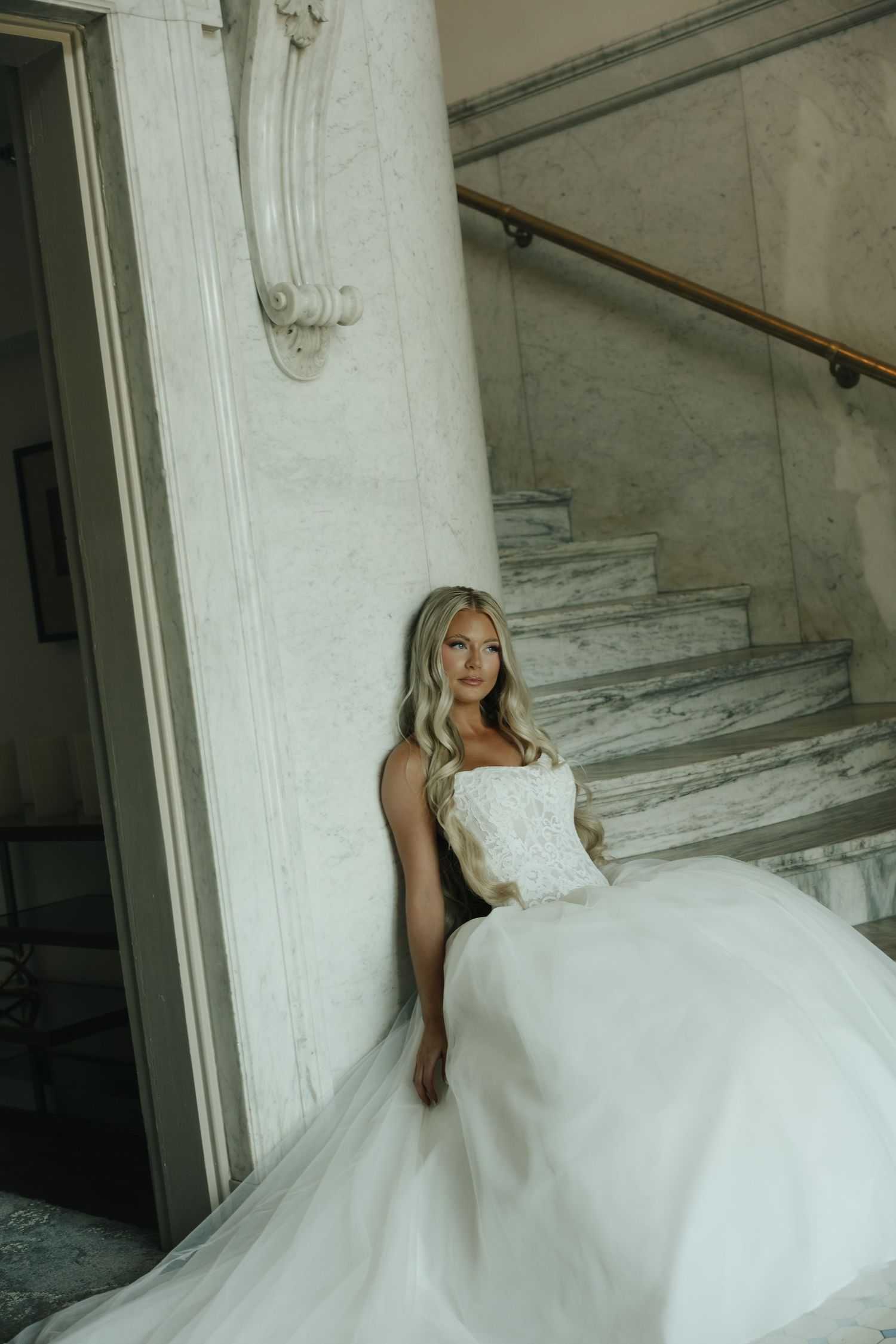 Bride in white ball gown with lace bodice leaning against marble column near elegant staircase.