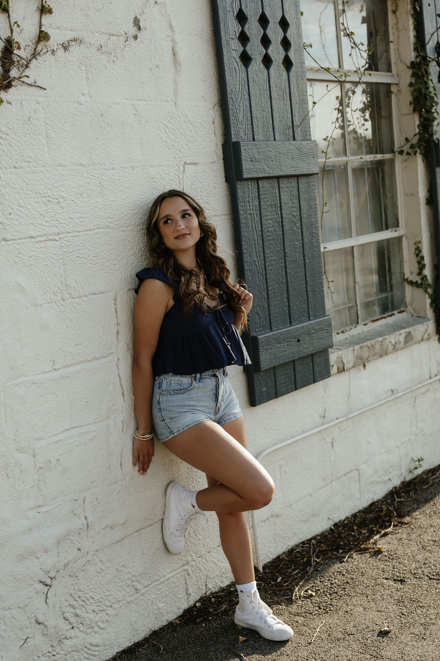 Young woman in denim shorts and navy top leans against white brick wall near vintage window with shutters.