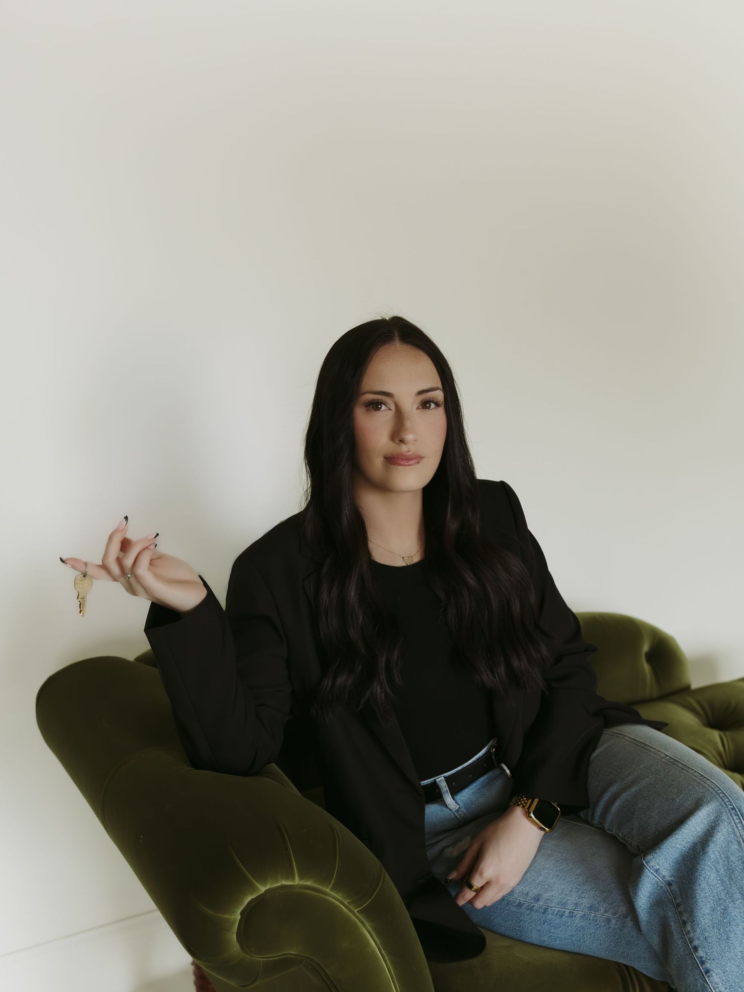Woman in black blazer and jeans sitting on green velvet sofa, holding jewelry, against white wall.