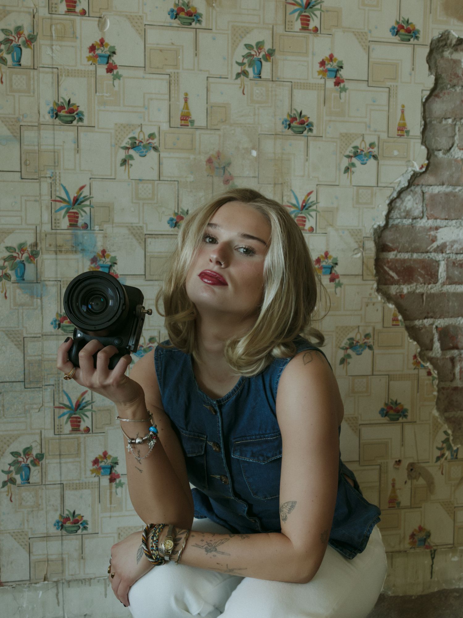 Blonde woman in denim top holds camera, sitting against vintage floral wallpaper with exposed brick, tattoos visible.