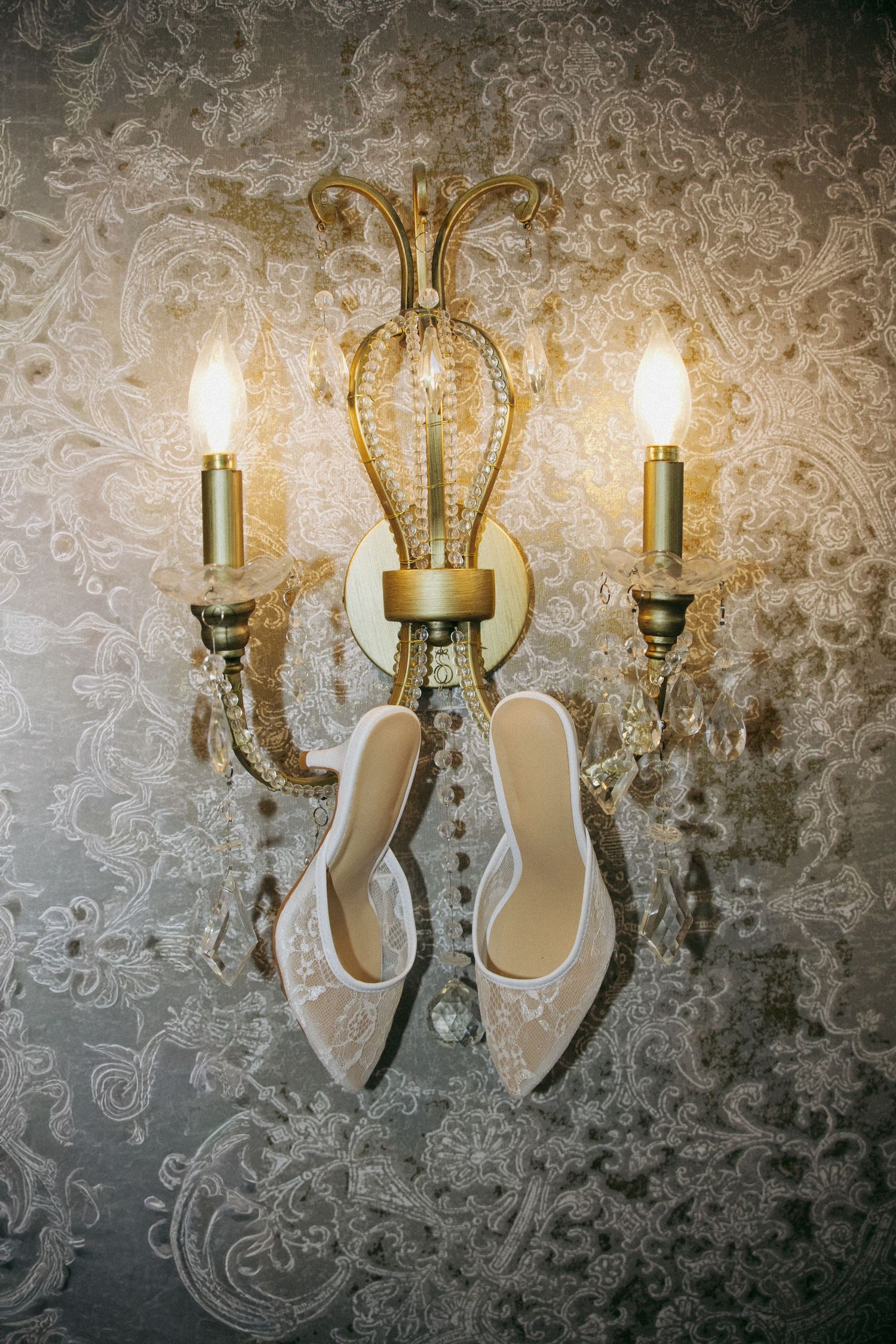 White lace bridal heels hanging on an ornate gold crystal wall sconce with lit candles against floral wallpaper.