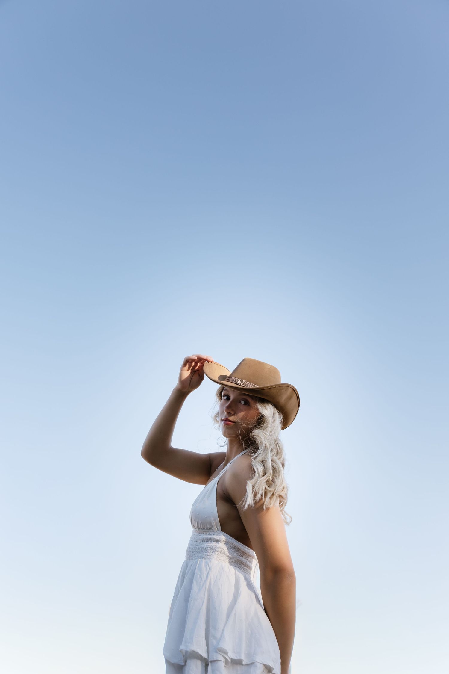 Blonde woman in white dress and tan cowboy hat posing against clear blue sky on a sunny day.