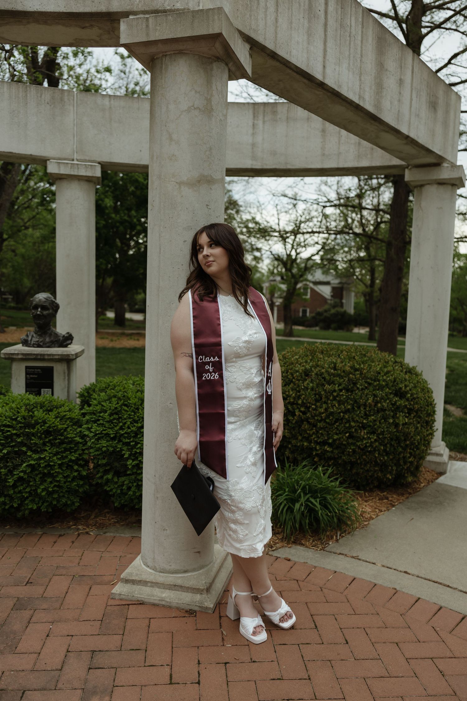 A young woman in a white dress and maroon graduation stole poses by stone columns on a brick path.