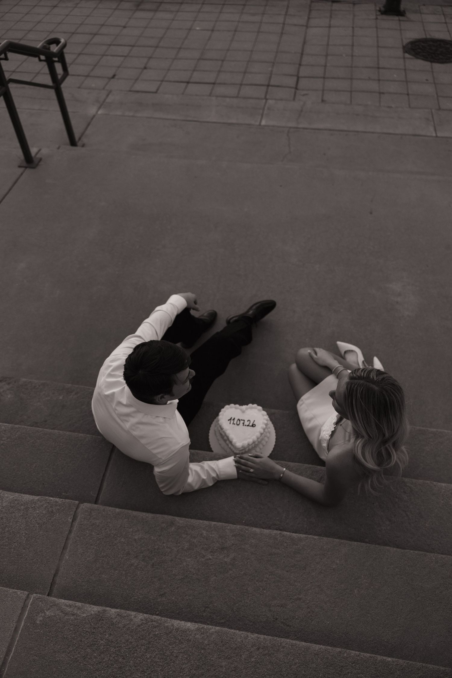 Aerial view of couple sitting on steps holding a cake, black and white wedding photo.