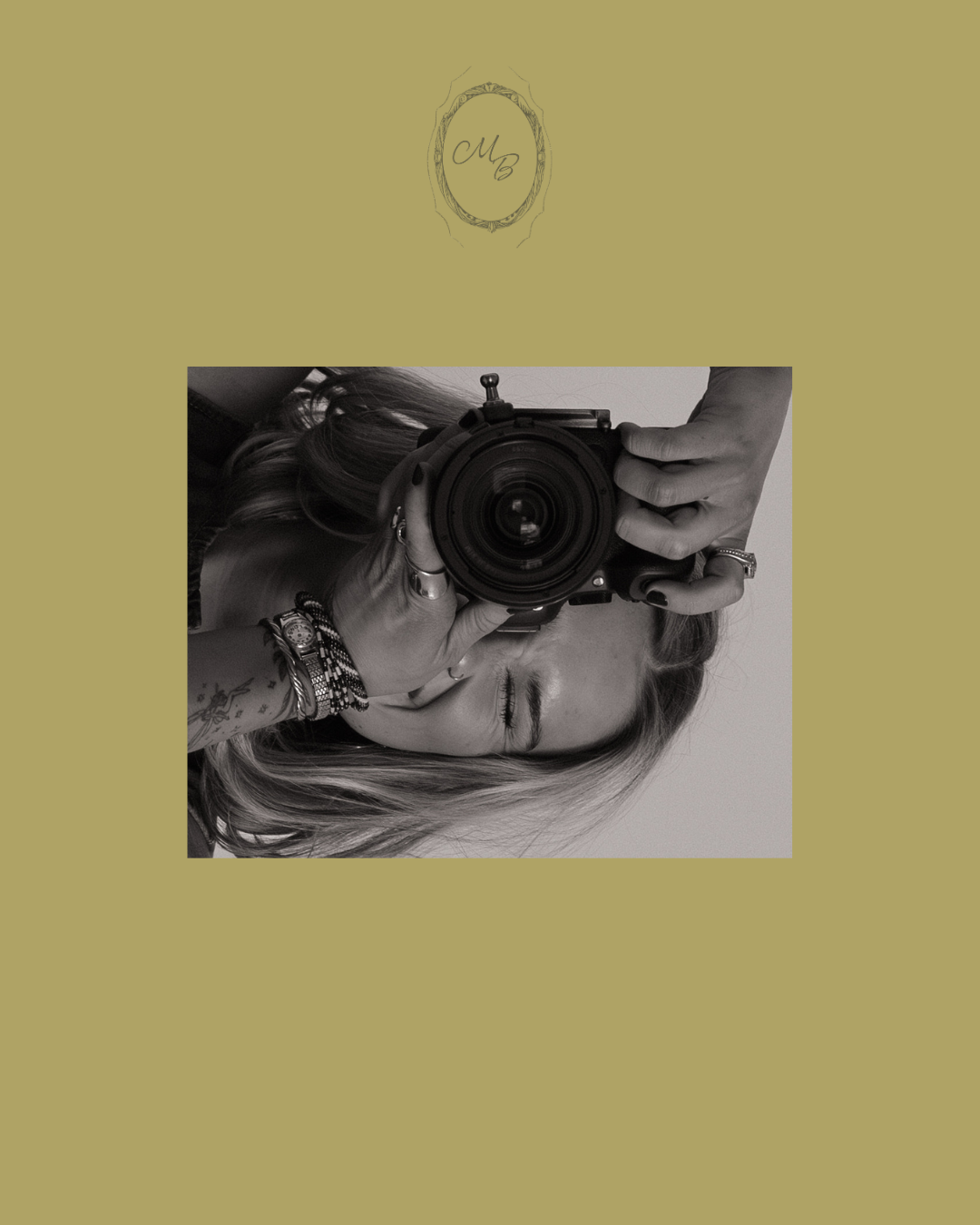 A woman holds a large camera up to her face, wearing stacked bracelets and rings, in a sepia-toned portrait.