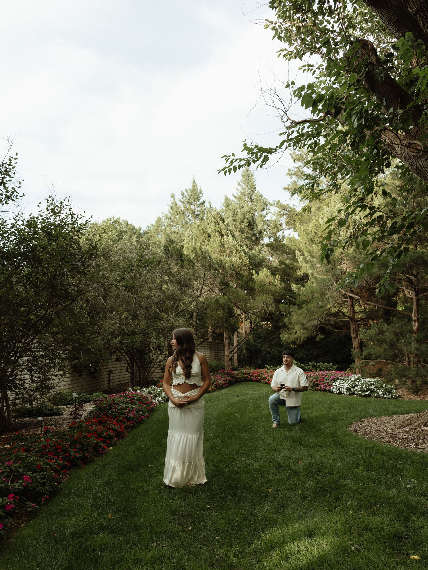 Man kneeling on grass proposing to woman in white dress in lush garden setting with colorful flowers and trees.