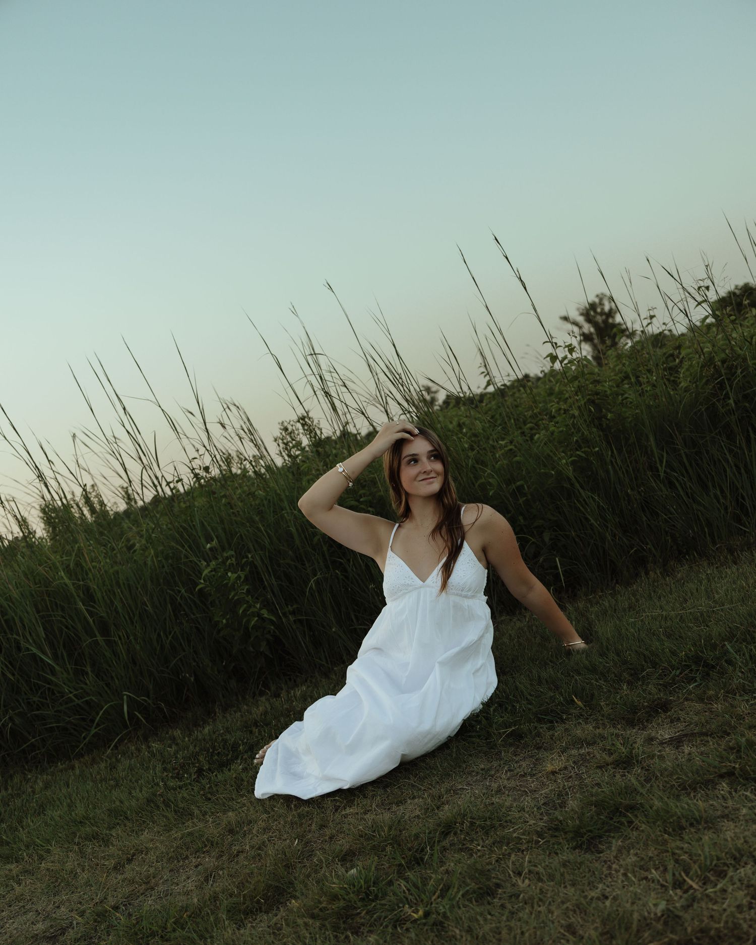 A smiling woman in a white sundress sits gracefully on green grass near tall reeds at dusk.