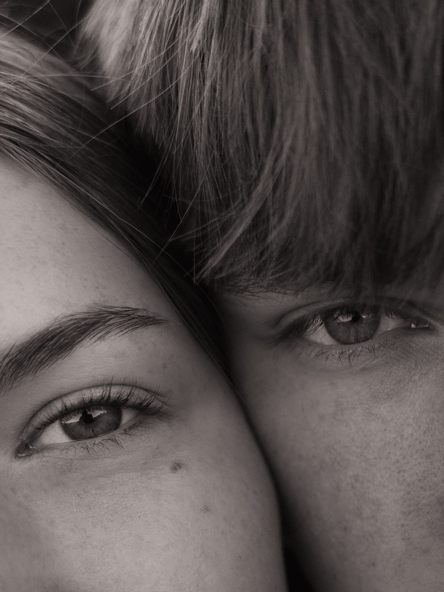 Black and white close-up portrait of two people with foreheads together, showing expressive eyes and long lashes.