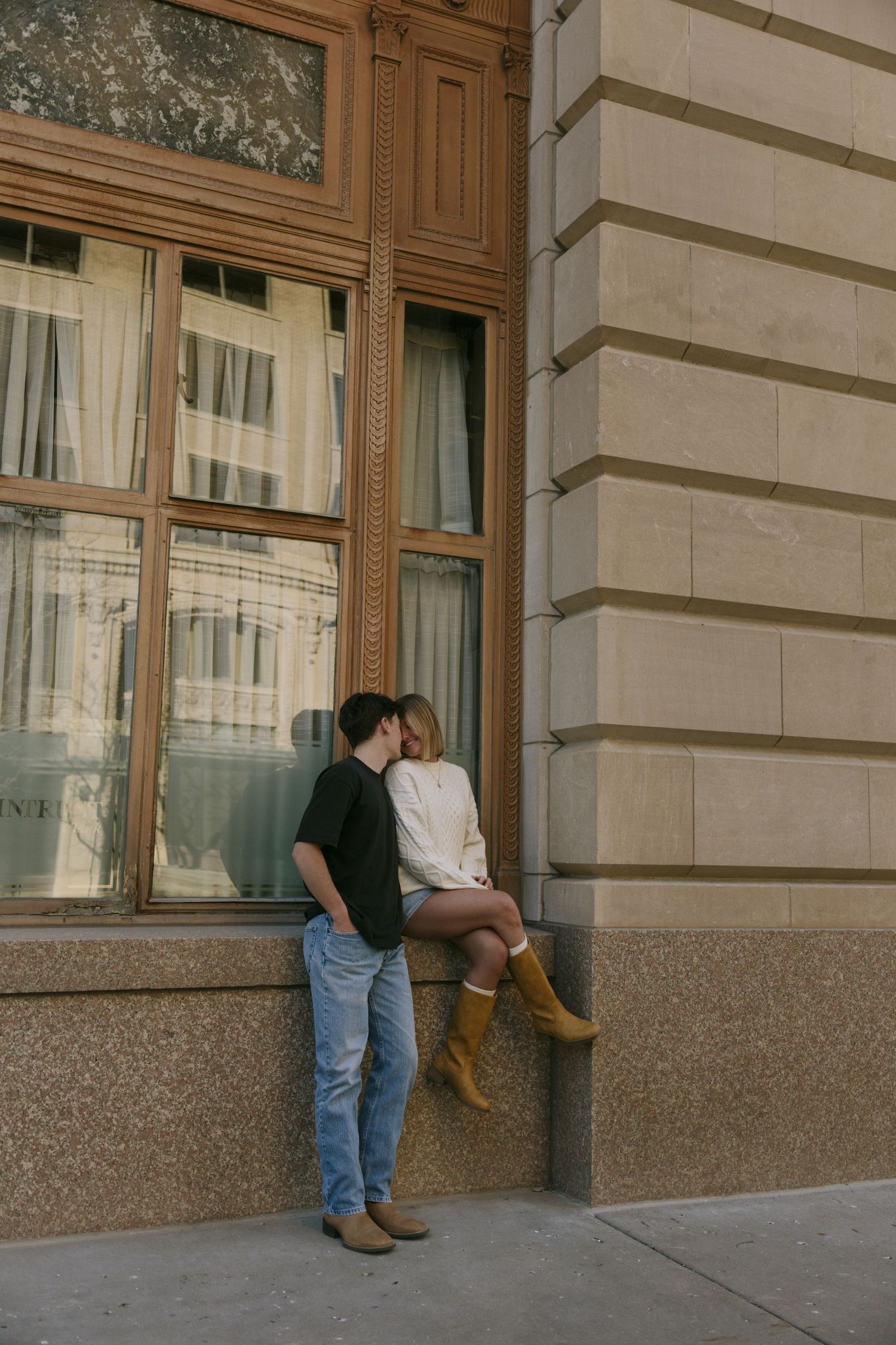 Couple sharing a kiss on a building ledge, woman in white sweater and tan boots, man in black tee and jeans.