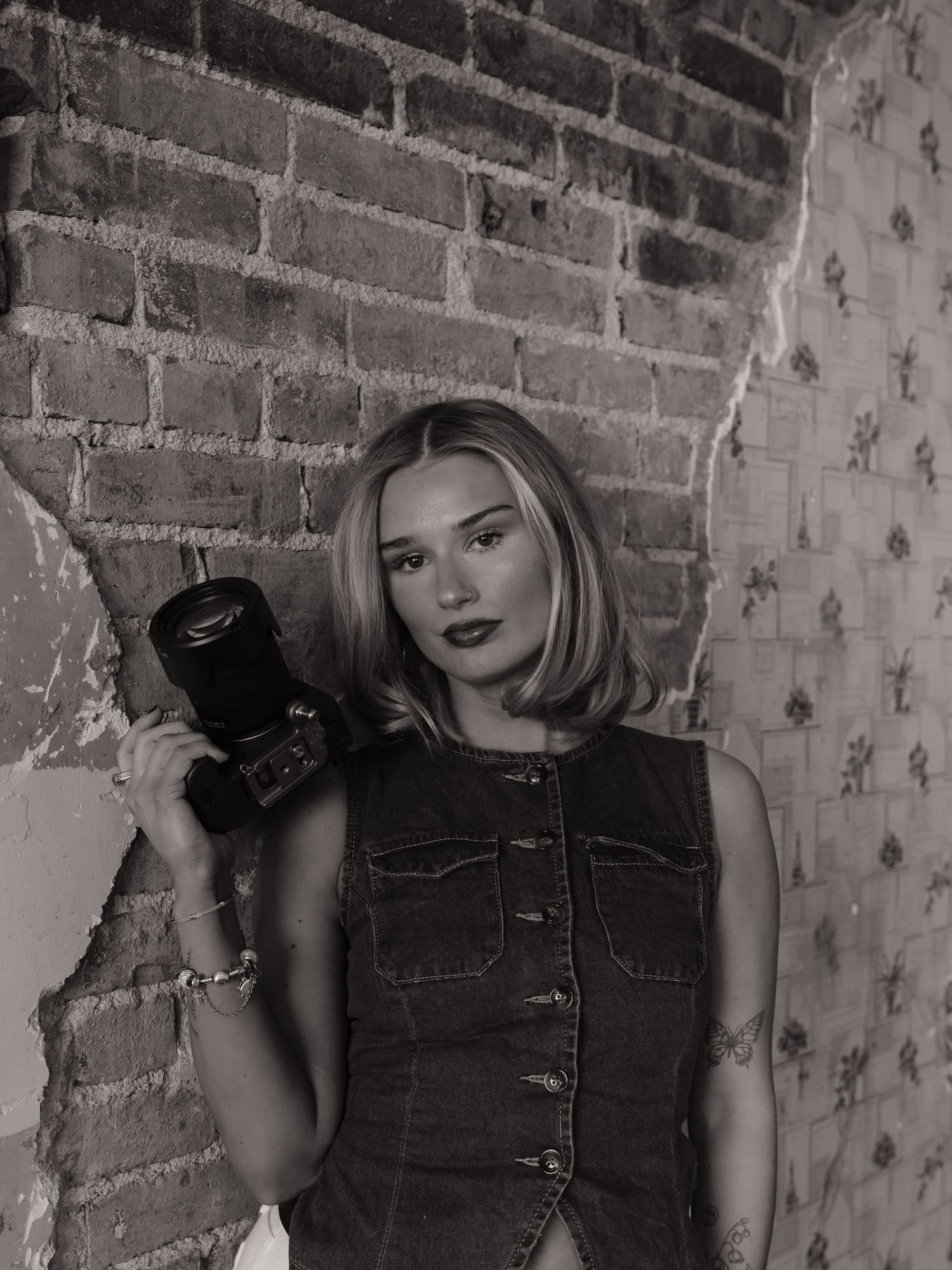 Black and white photo of a blonde woman in denim dress holding a camera against a brick wall.