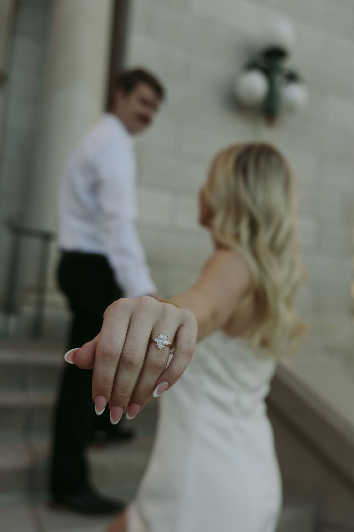 Bride shows off her engagement ring while holding hands with groom on steps during wedding photo session.