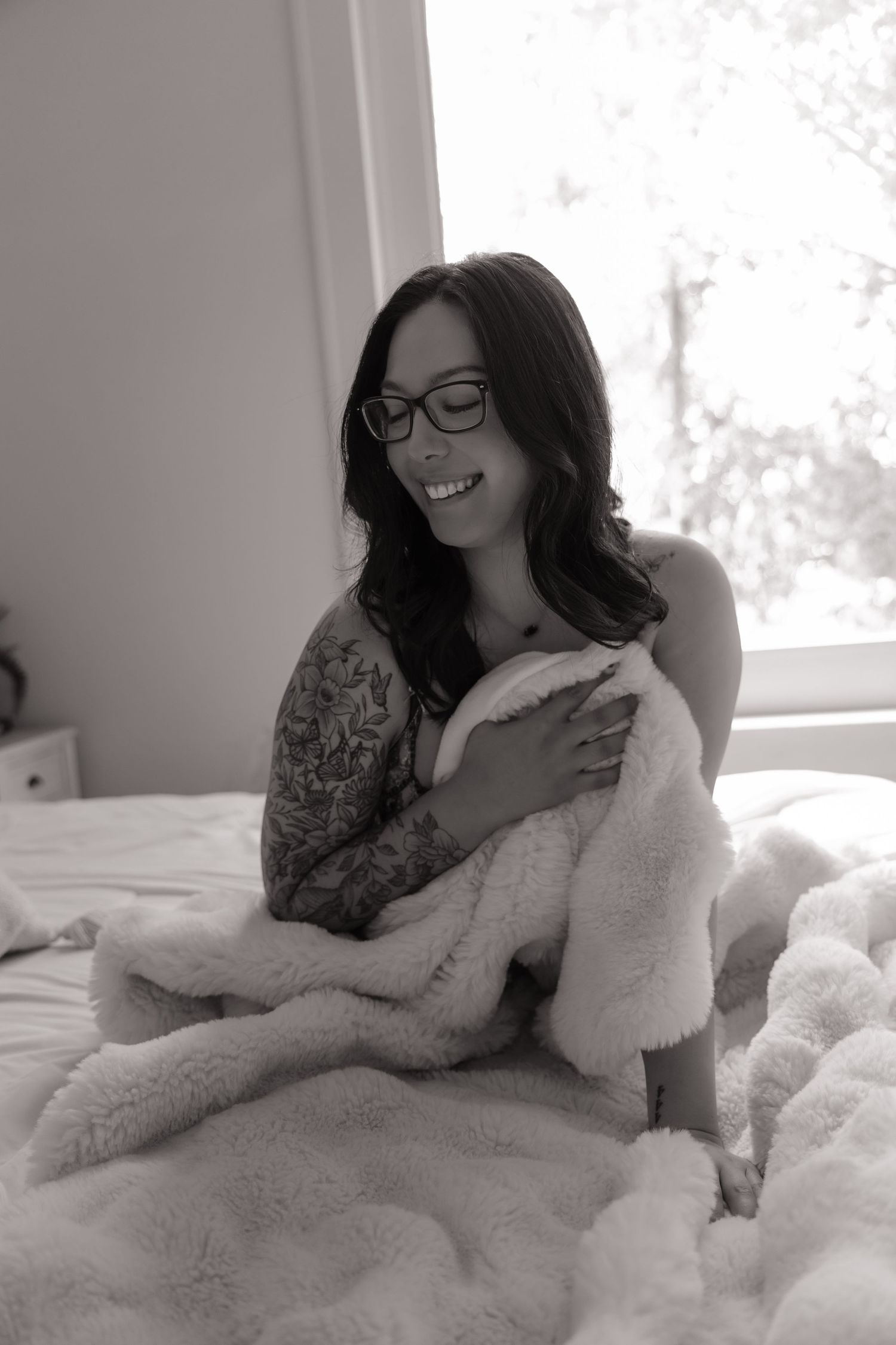 Smiling woman with glasses and tattoos wrapped in fluffy blanket sitting on bed, black and white photo.