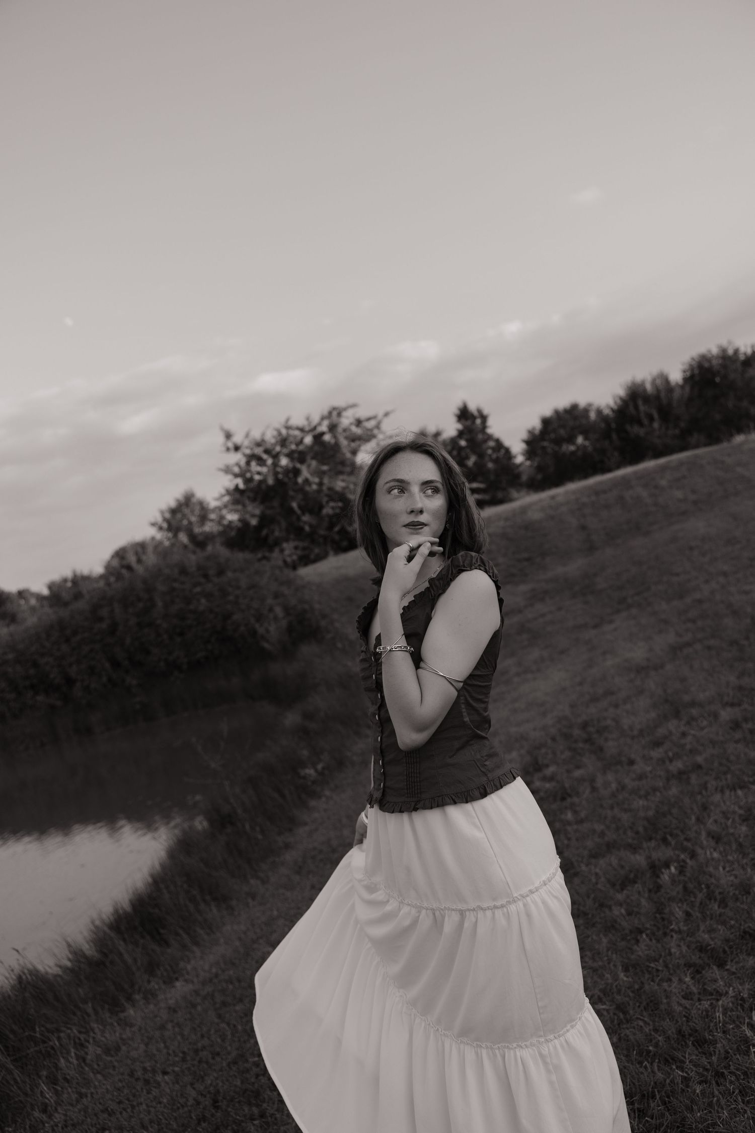 Black and white photo of a woman in a white maxi skirt and vest standing in a field near a pond at dusk.