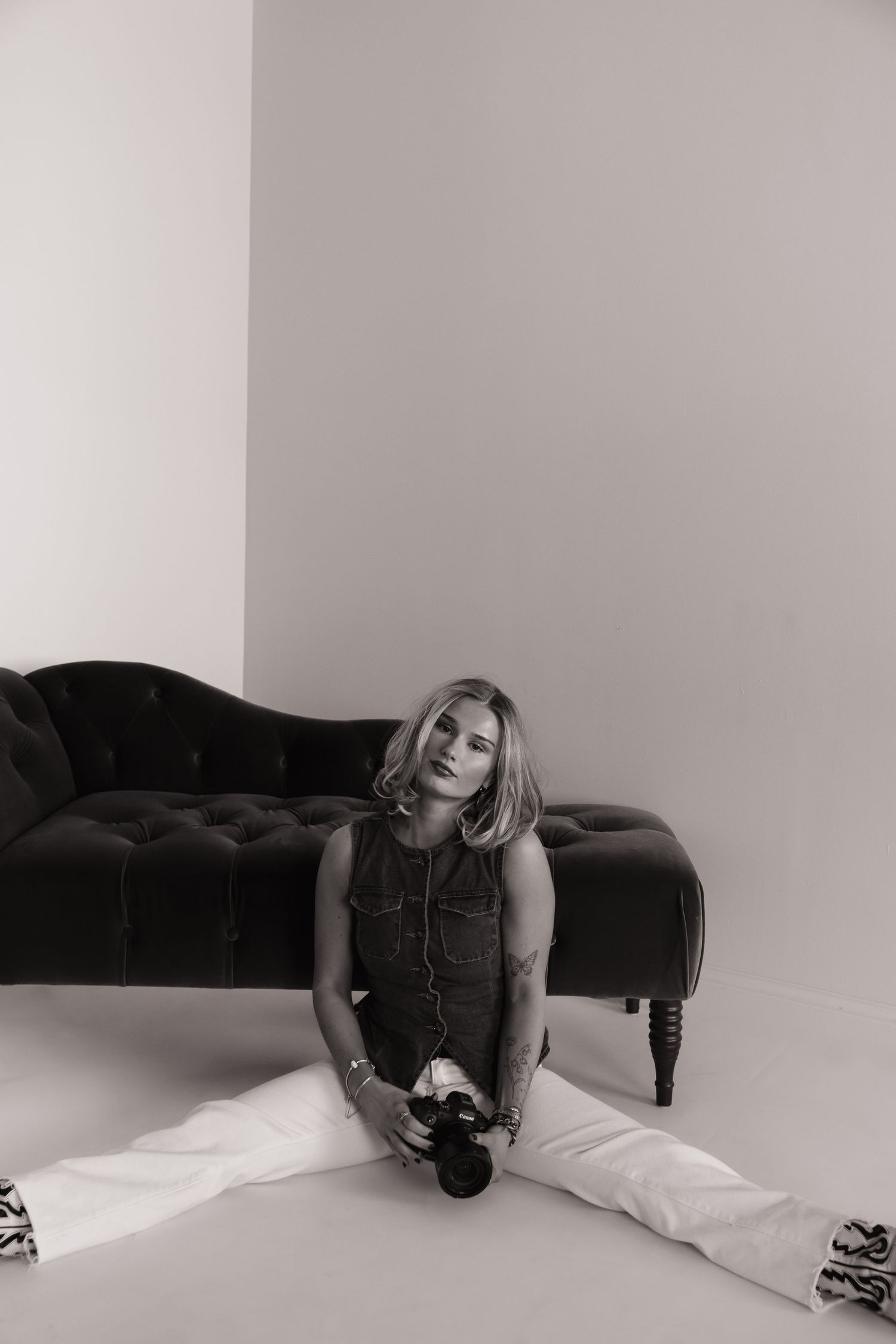 Black and white photo of a blonde woman sitting on floor holding a camera beside a tufted velvet sofa.