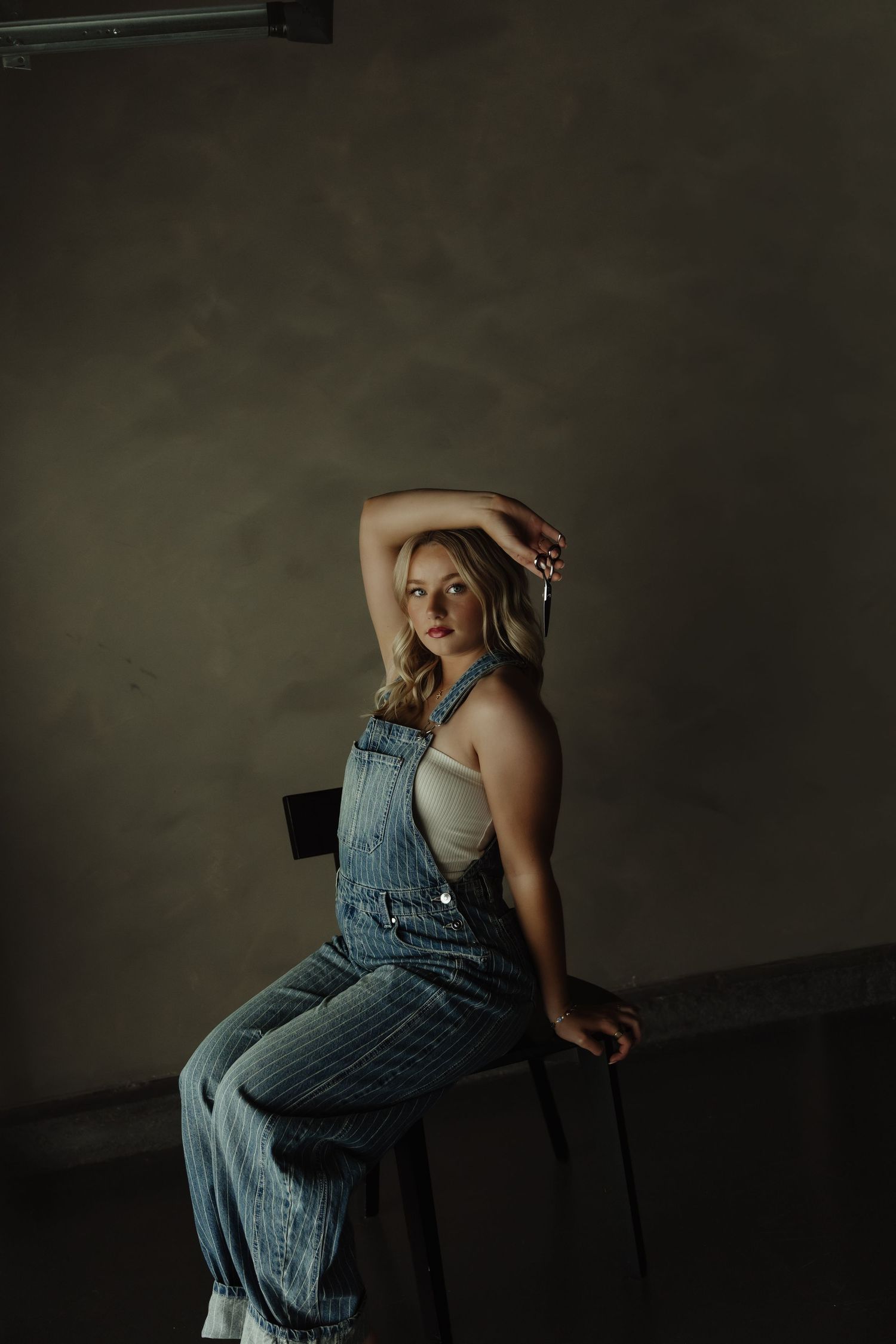 Young woman in denim overalls poses gracefully on a stool against a moody studio backdrop.