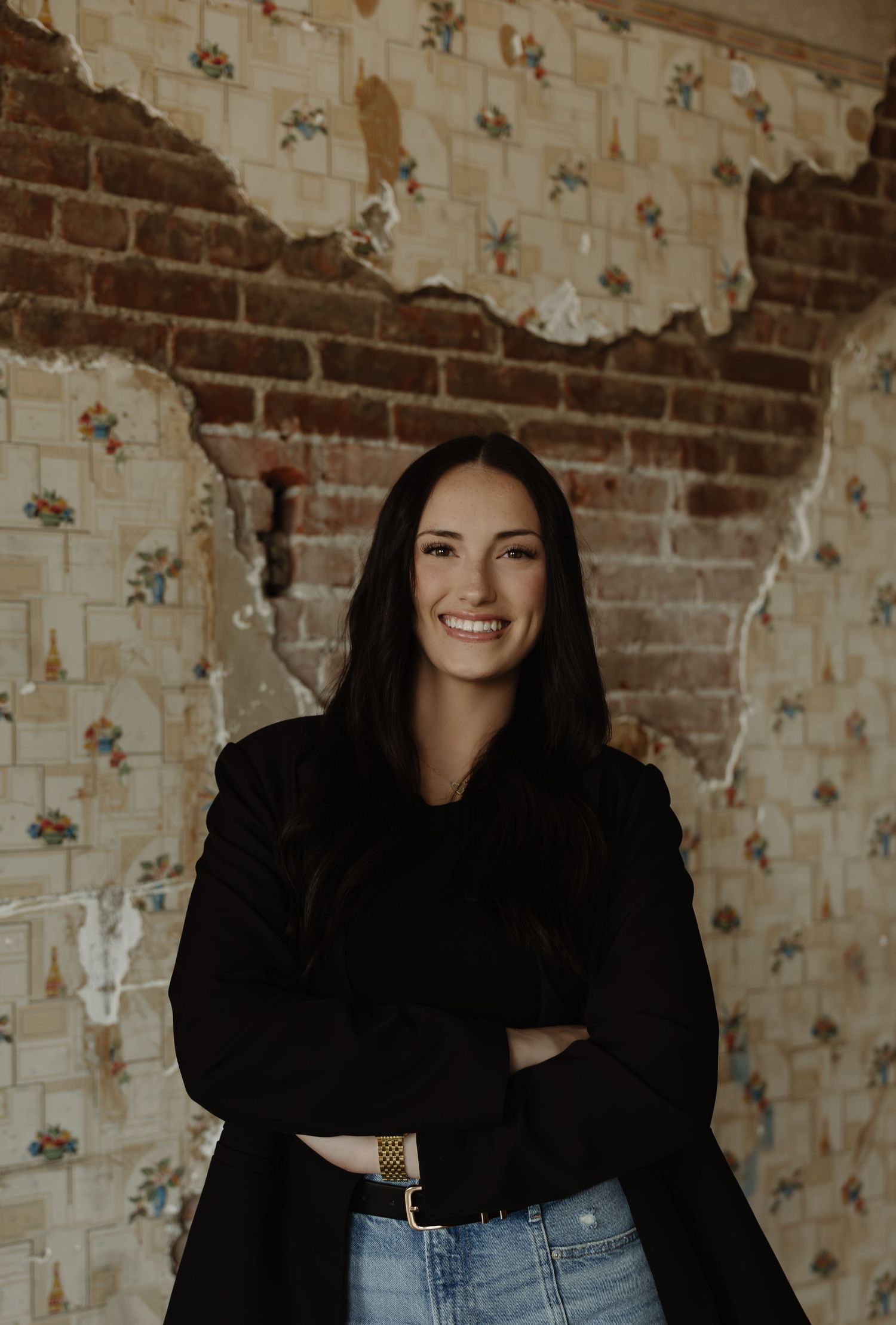 Smiling woman with dark hair, arms crossed, wearing black jacket and jeans, posed against rustic brick and floral wallpaper.