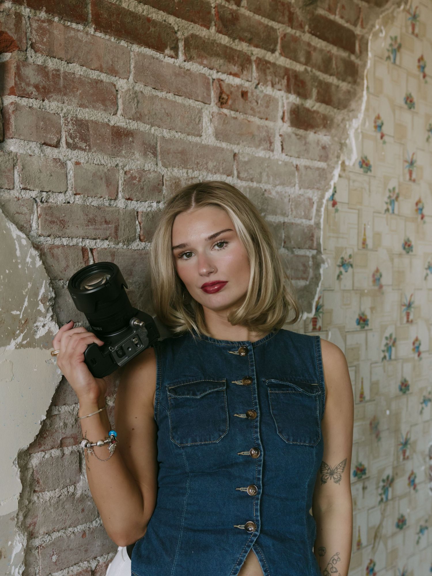 Blonde woman in denim dress holding a camera, posing against a rustic brick wall with vintage wallpaper.