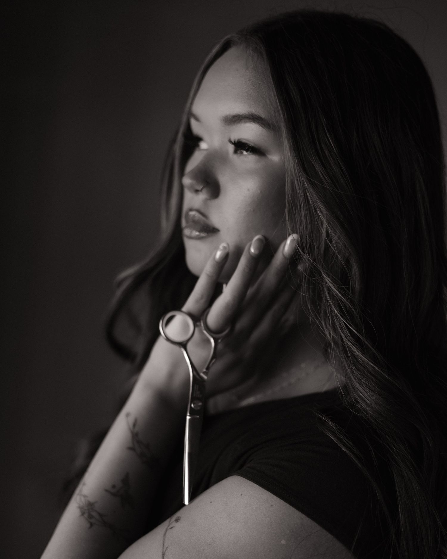 Black and white portrait of a young woman with long hair, jewelry, and tattoos posing thoughtfully.