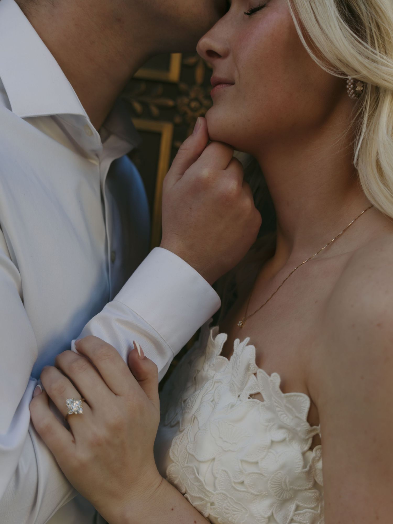 Bride in white feather gown and groom in dress shirt share an intimate moment, showcasing her diamond engagement ring.