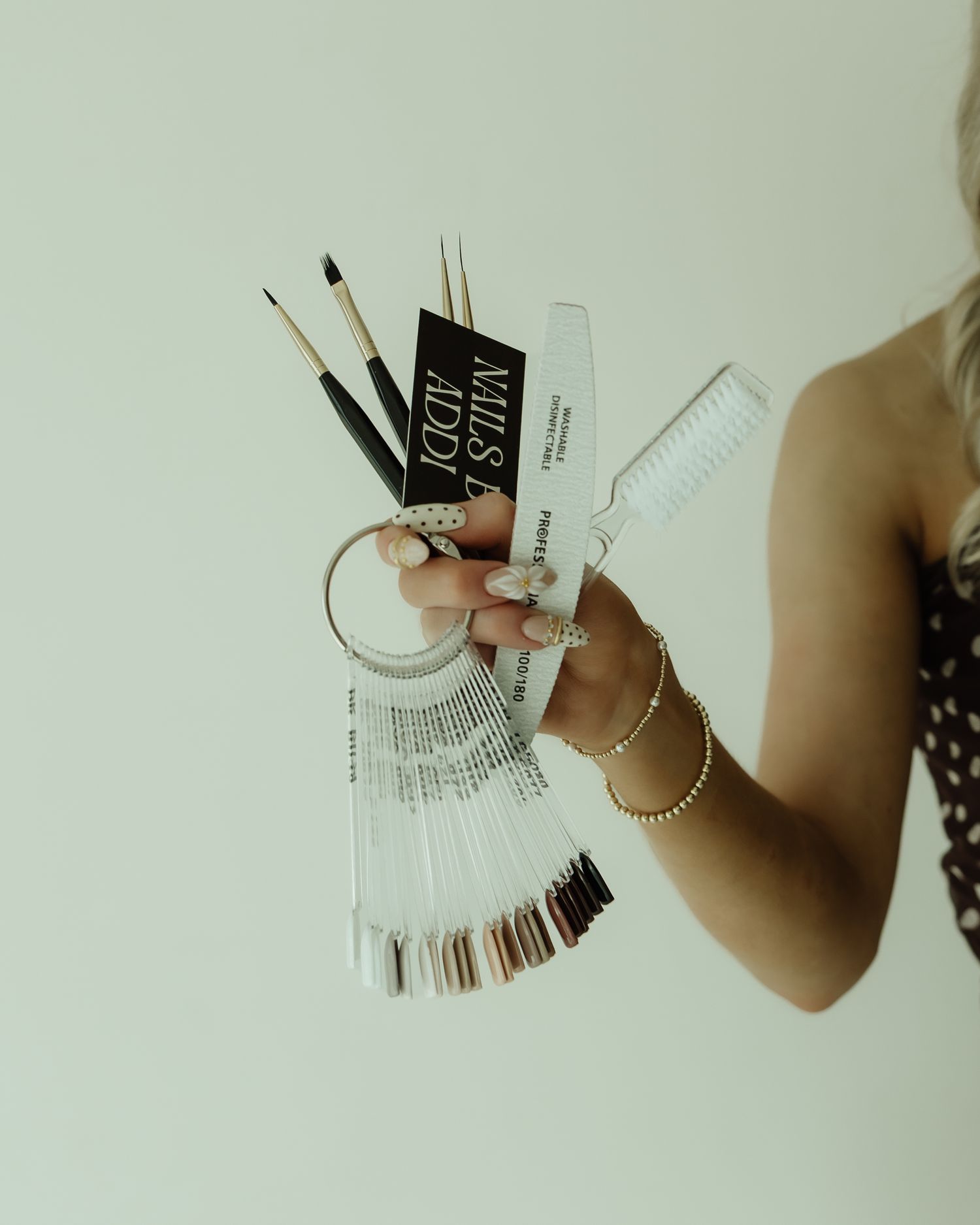 A nail technician holds nail art brushes, color swatches, and tools, showcasing manicured nails with gold bracelets.