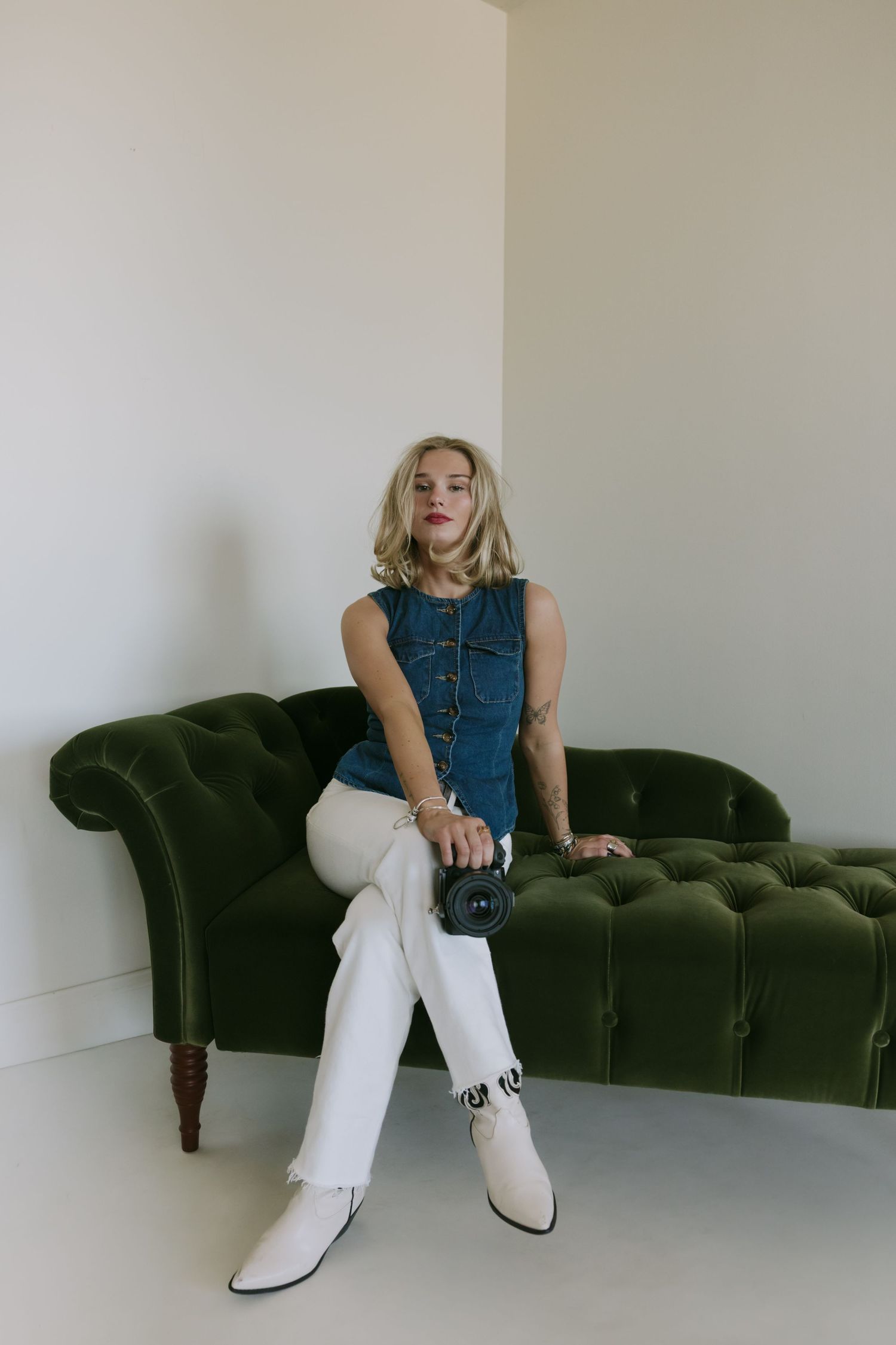 Blonde woman in denim vest and white pants sitting on green velvet tufted sofa holding a camera.