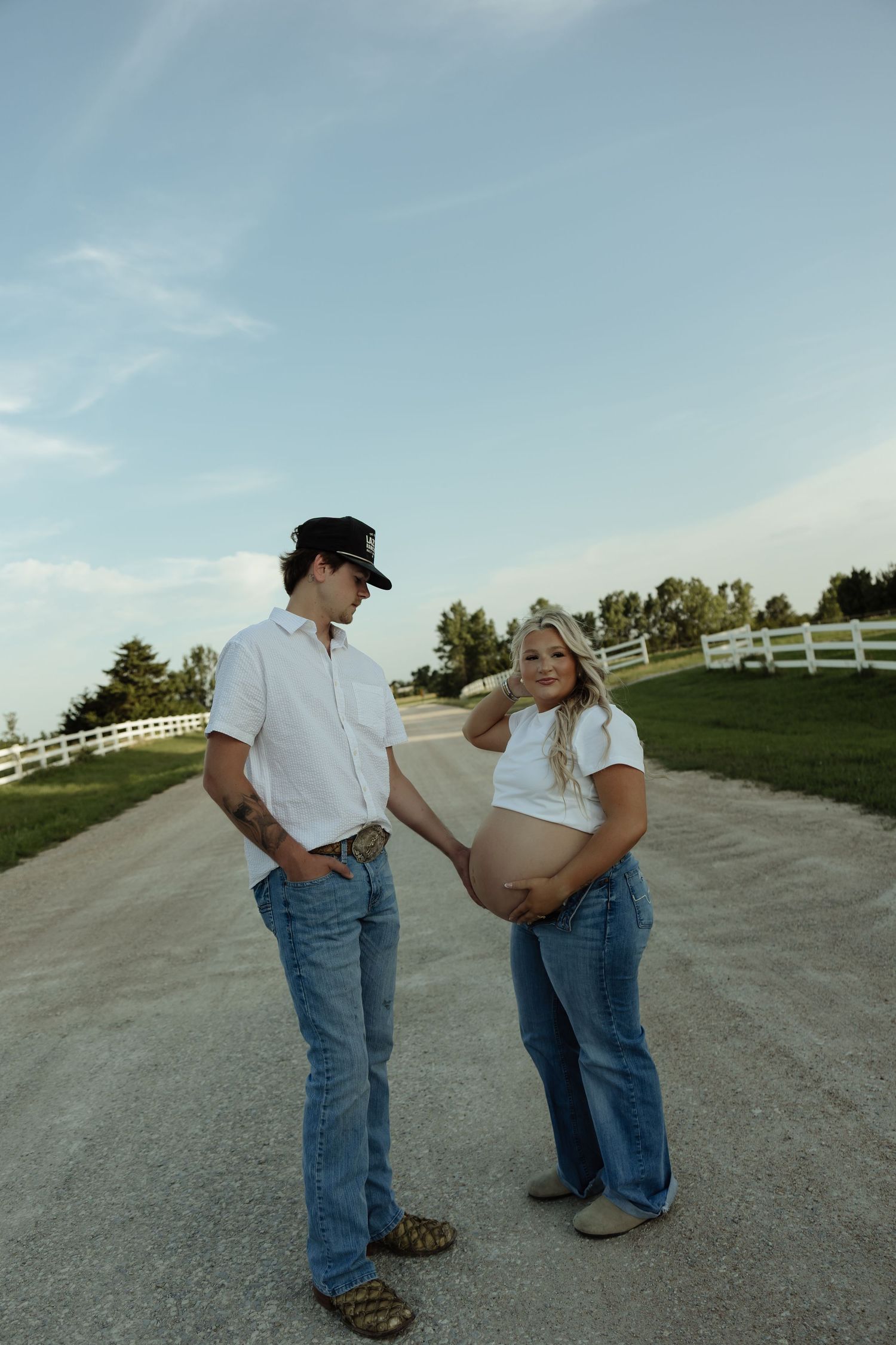 Pregnant couple in matching white shirts and jeans pose on a rural gravel road with white fence backdrop.