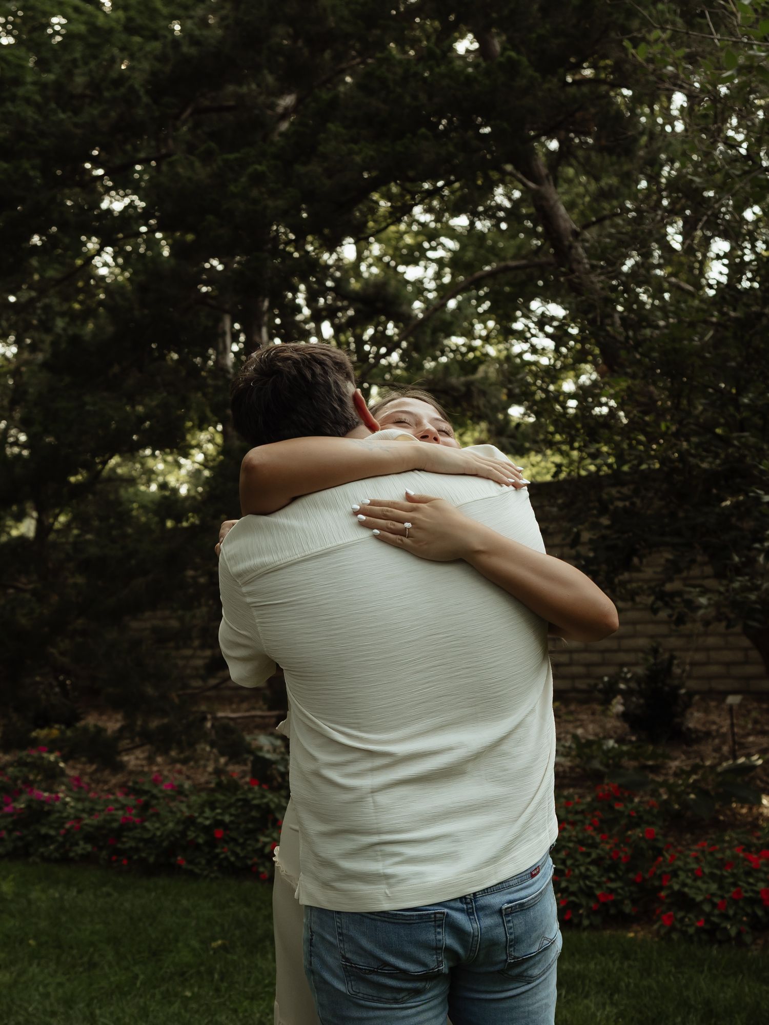 Couple sharing a warm embrace in a lush garden with blooming flowers and green trees in the background.
