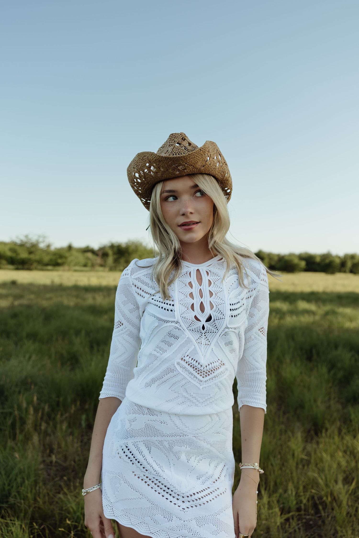 Blonde woman in white lace dress and brown cowboy hat standing in a green field under a clear blue sky.