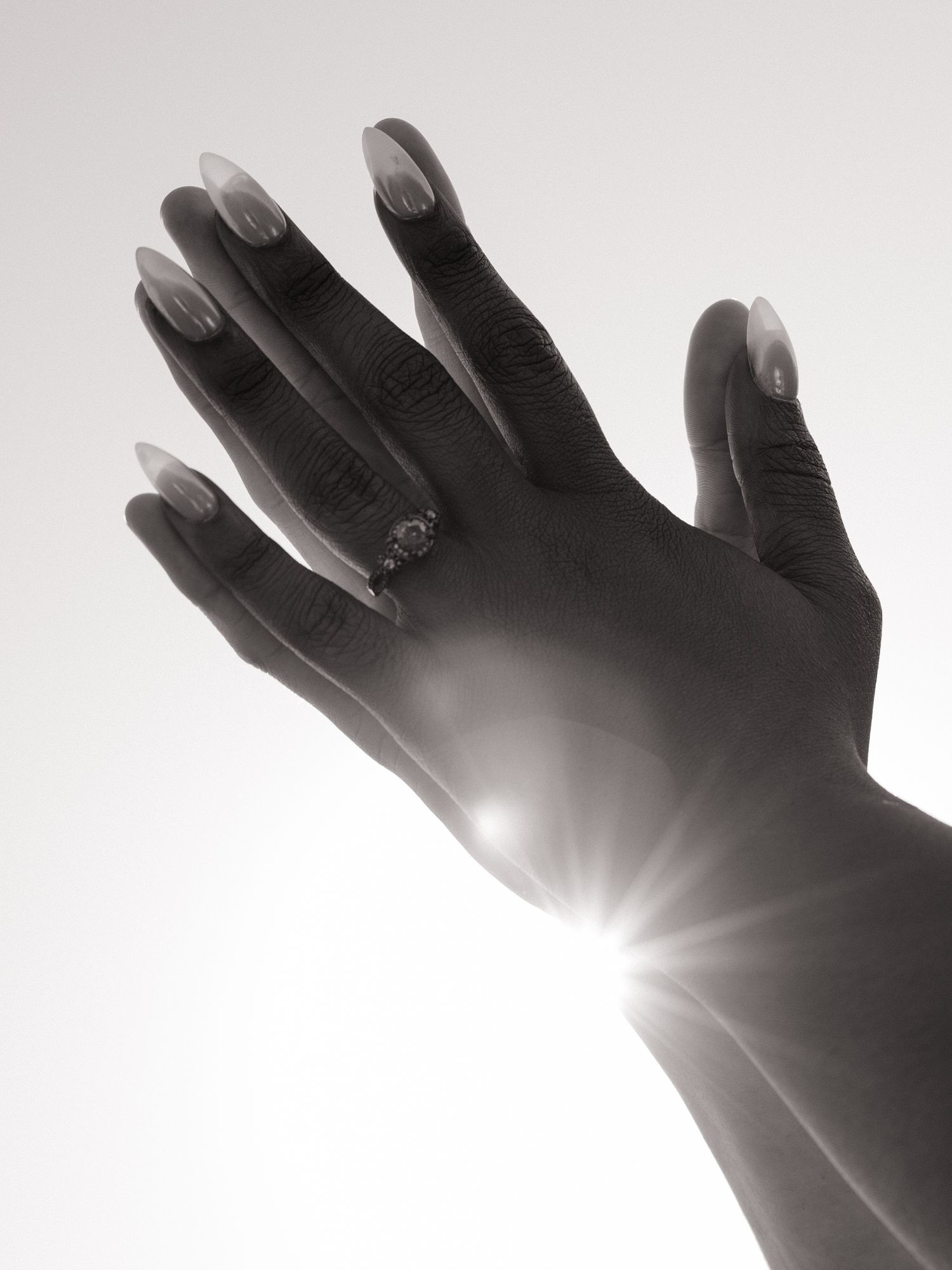 Black and white photo of a hand with long nails and a ring, sunlight bursting between fingers.