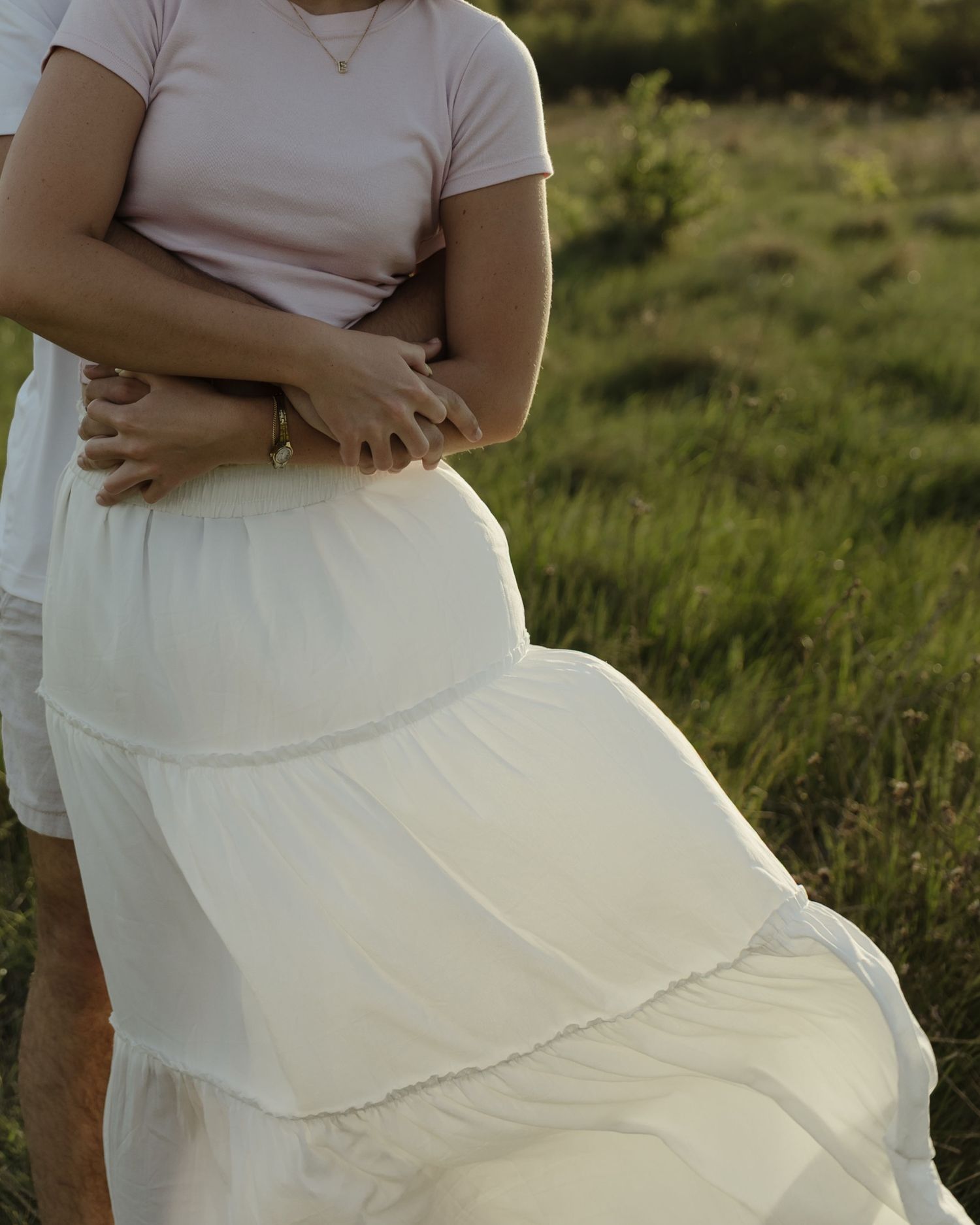 Couple embracing in a field, woman wearing a white tiered maxi skirt and pink top at golden hour.