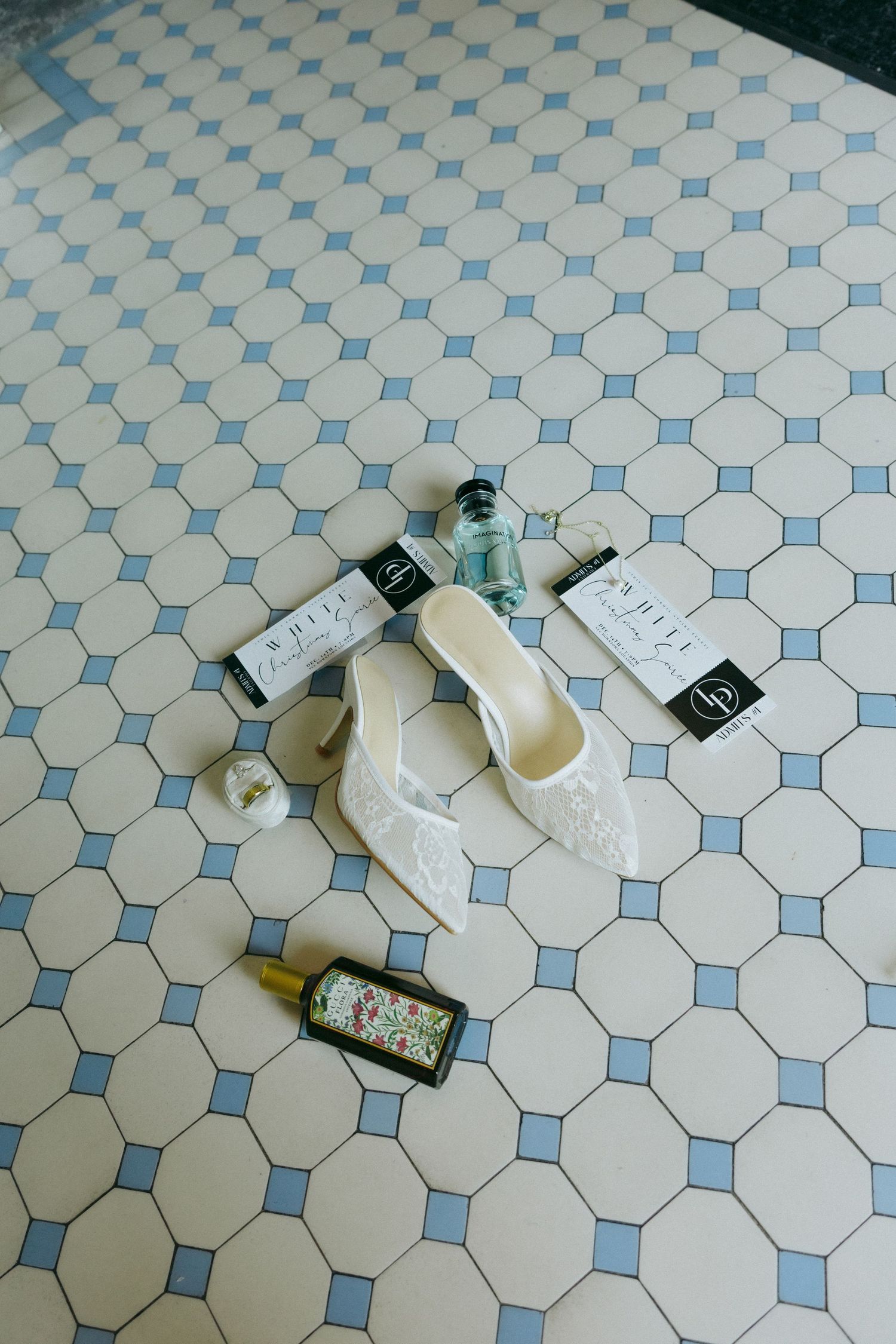 White lace bridal heels surrounded by perfume bottles and wedding cards on classic blue and cream hexagonal tile floor.