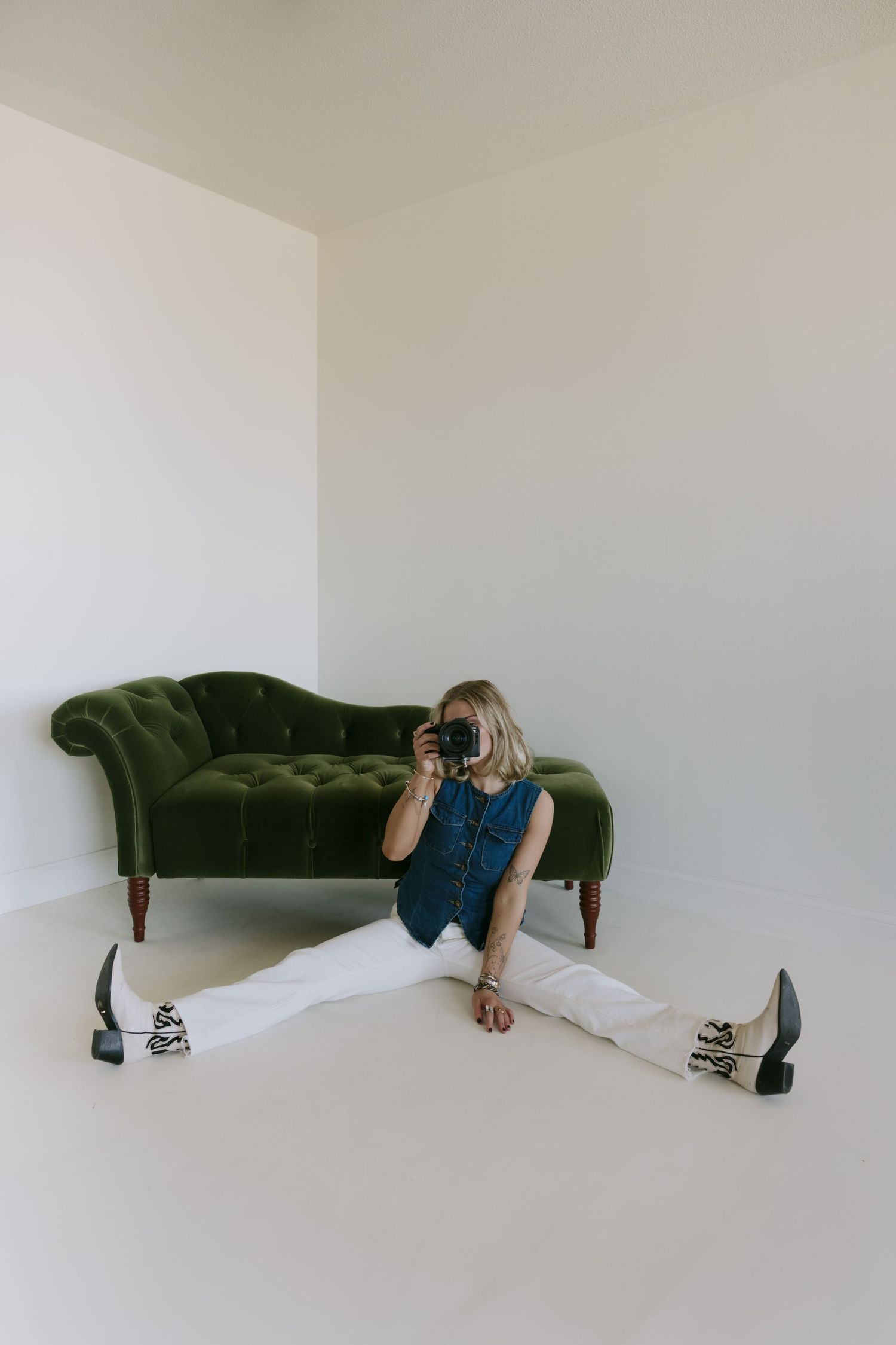 Woman sitting on floor in splits pose, holding camera, wearing white pants and blue top, near green velvet chaise lounge.