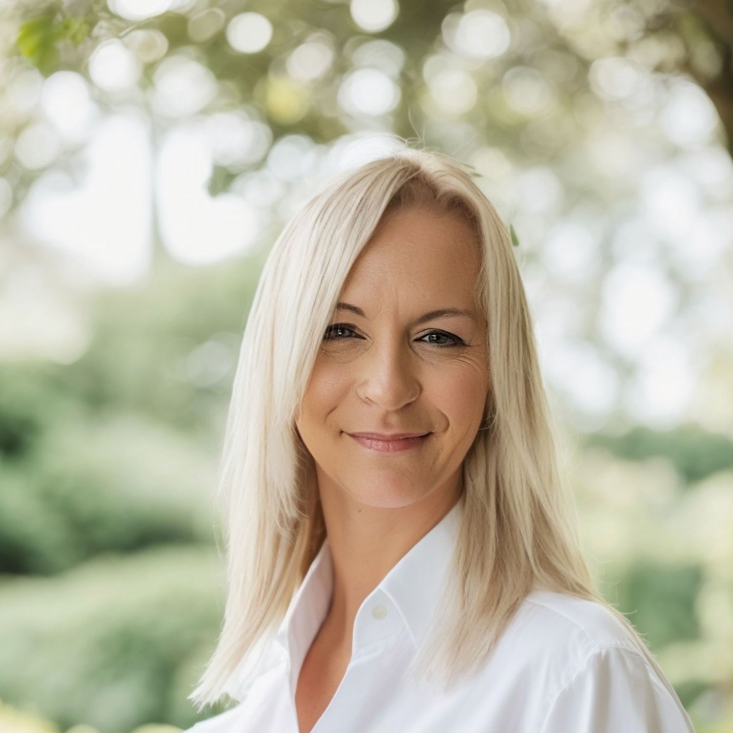Professional portrait of someone in a white button-down shirt against a soft outdoor bokeh background.