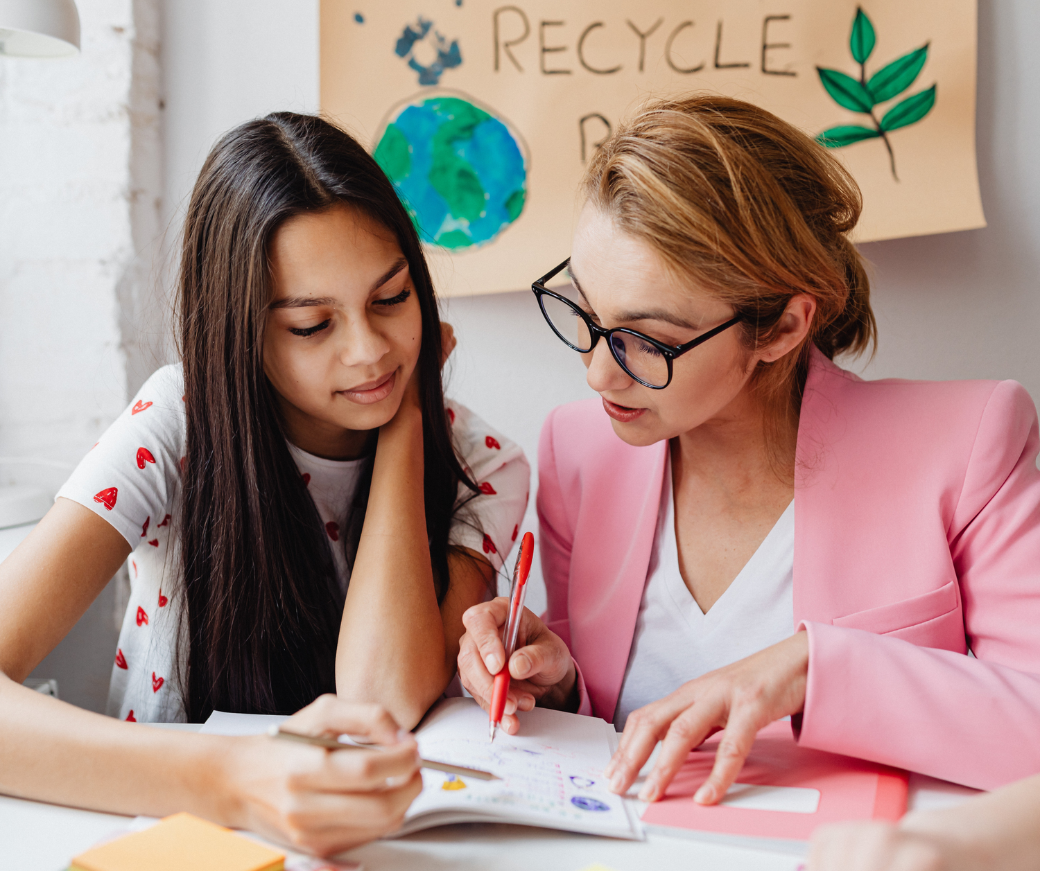Two students working together at a desk with recycling and environmental education materials displayed on the wall.