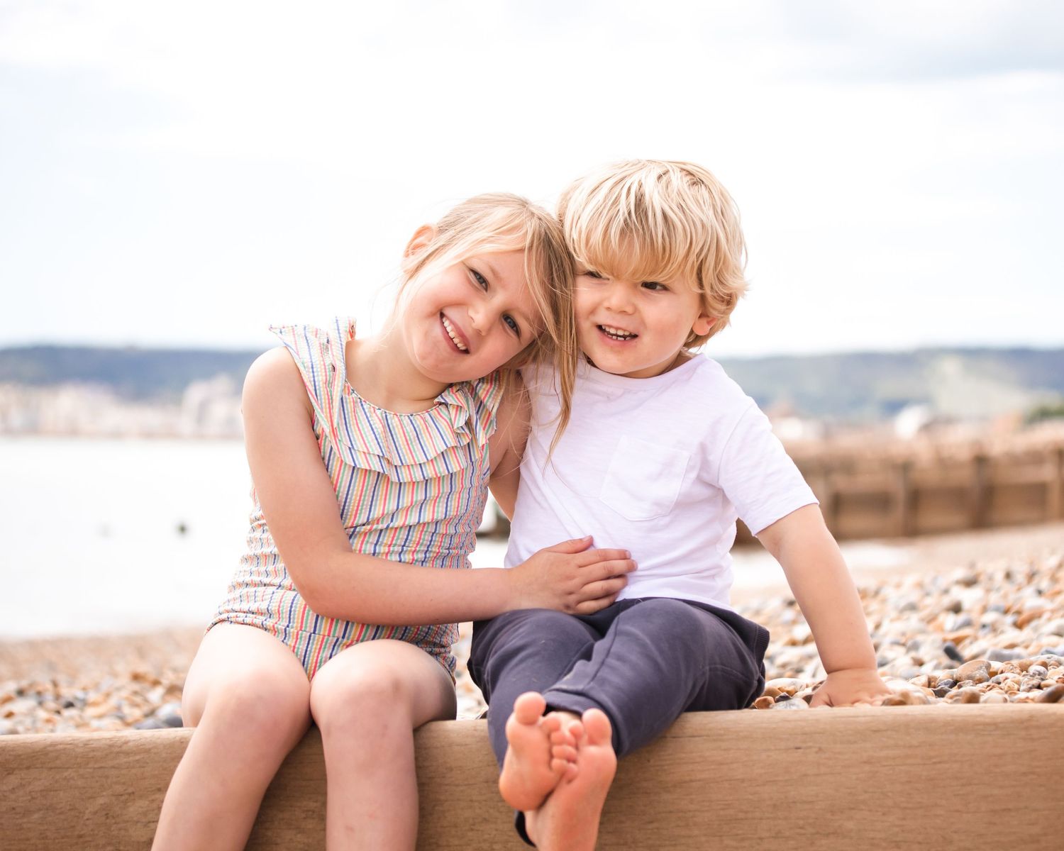 brother and sister sit on the eastbourne beach cuddling and smiling