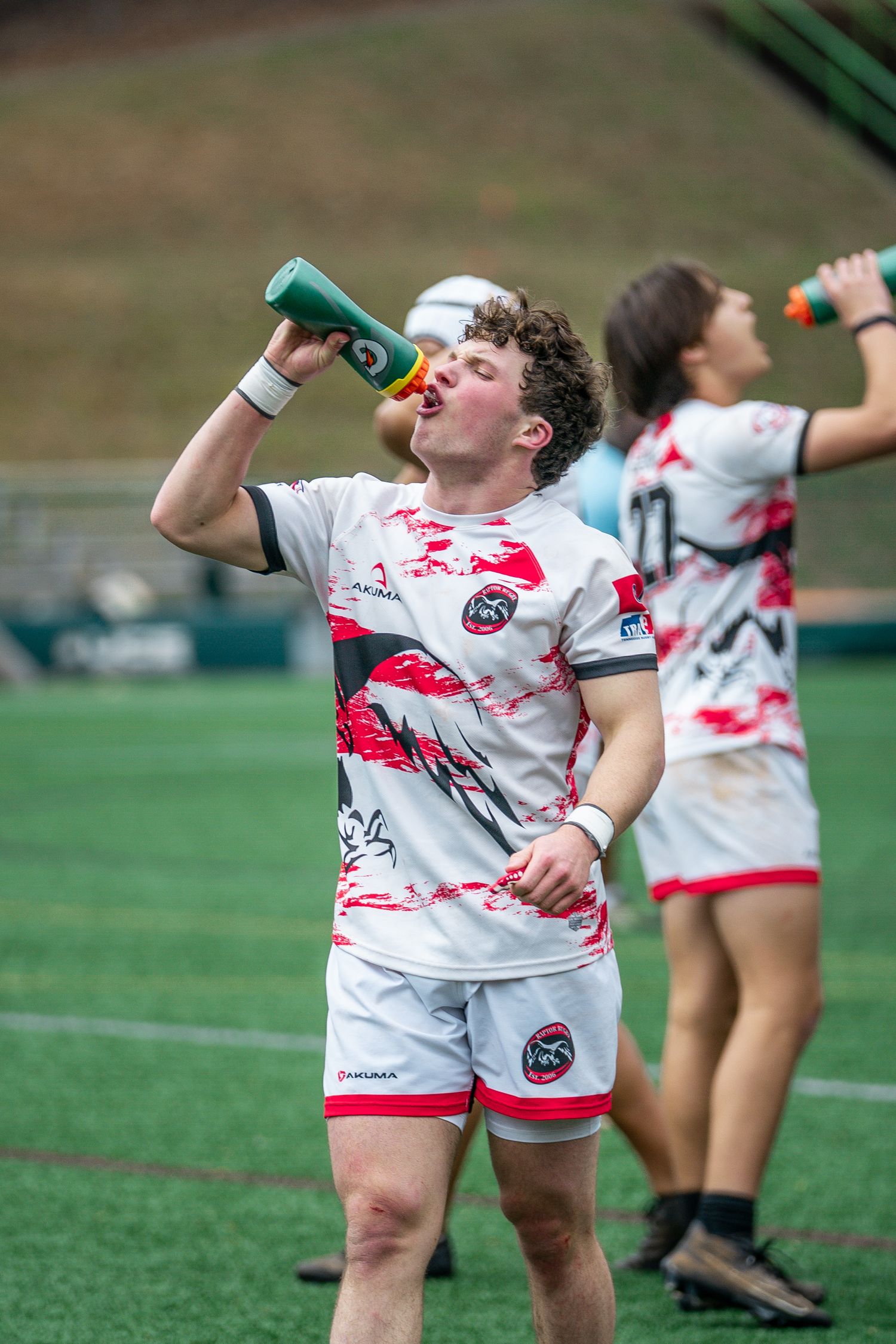 Rugby players in white and red uniforms hydrate during a match break on a grass field.