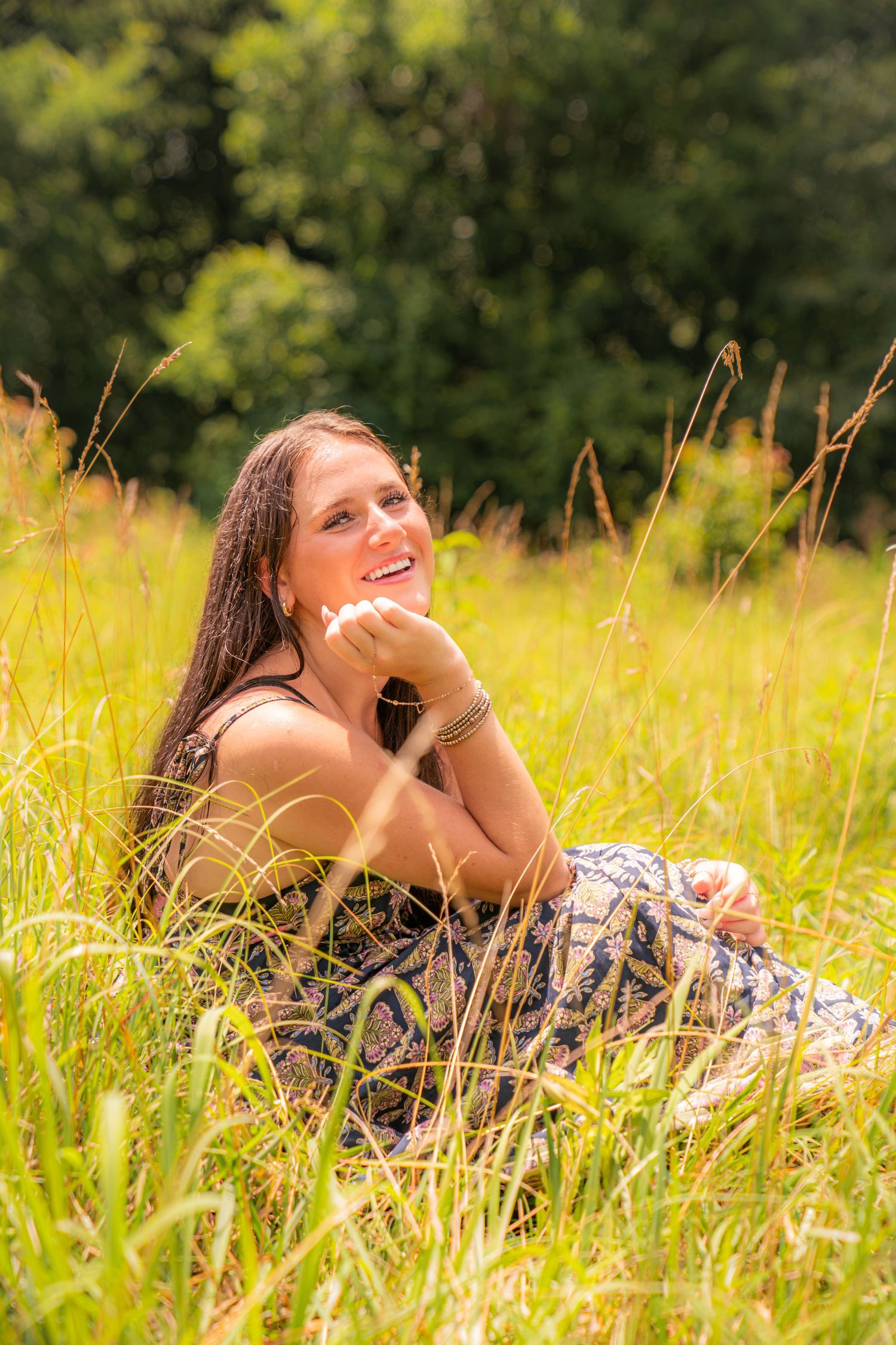 Young woman in floral dress sitting in tall grass field, smiling and looking up on a sunny day.