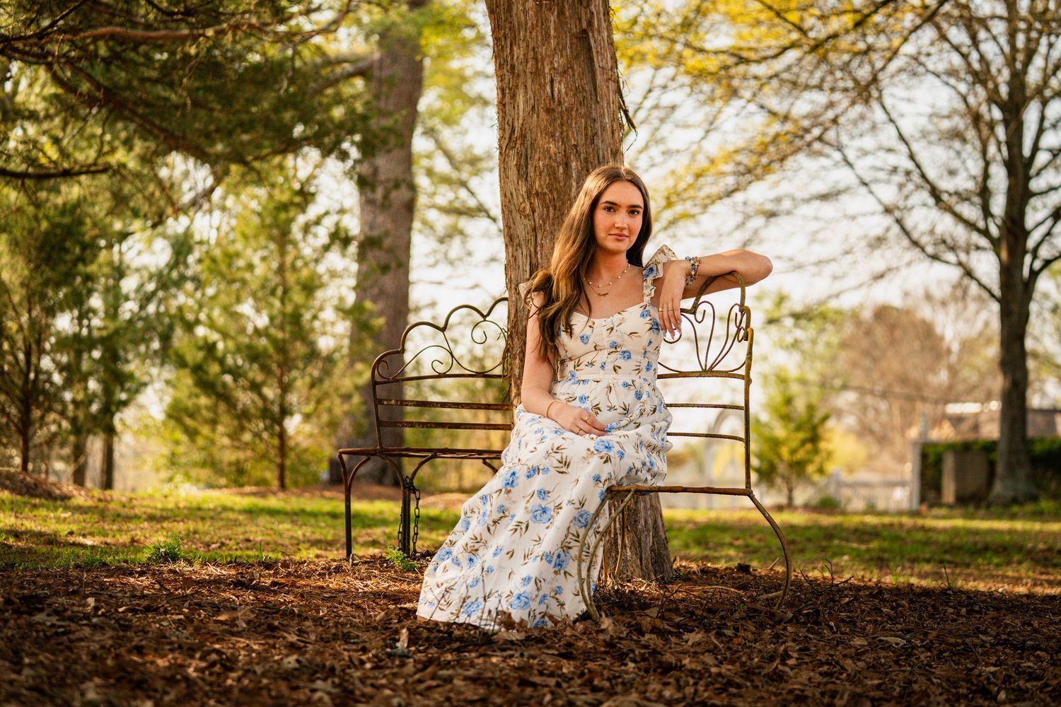Young woman in floral maxi dress sitting on ornate bench in autumn park with golden trees.