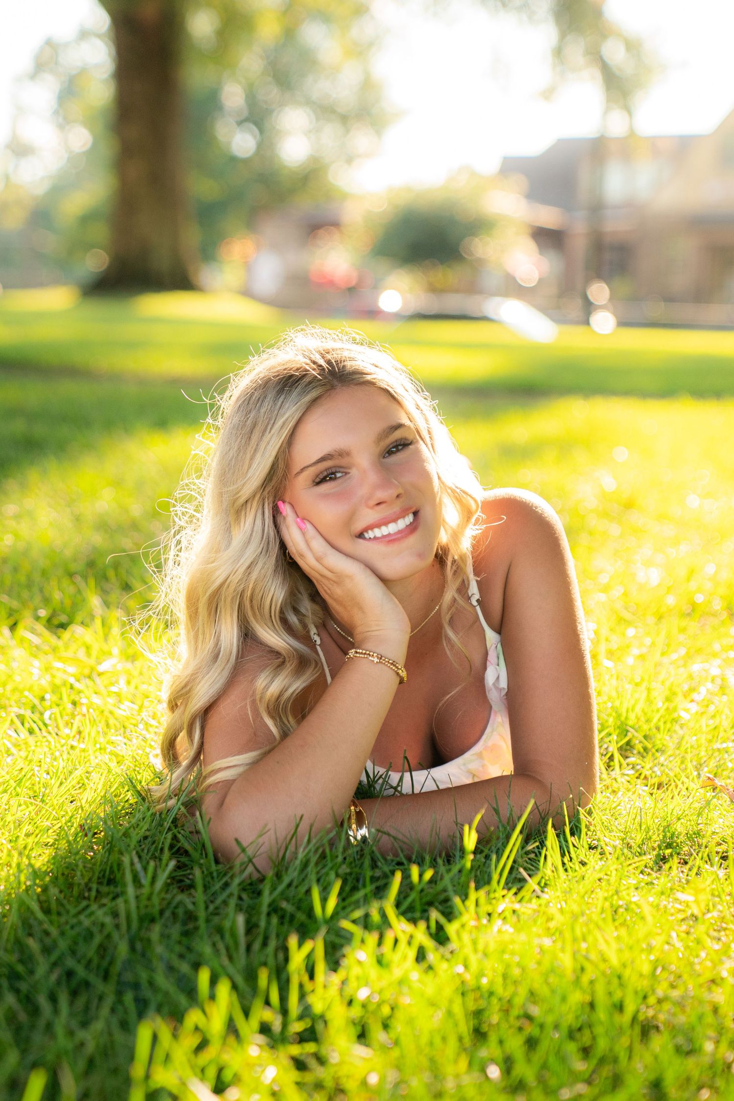 Smiling blonde woman lying on green grass in sunlight, resting chin on hand, in an outdoor park setting.
