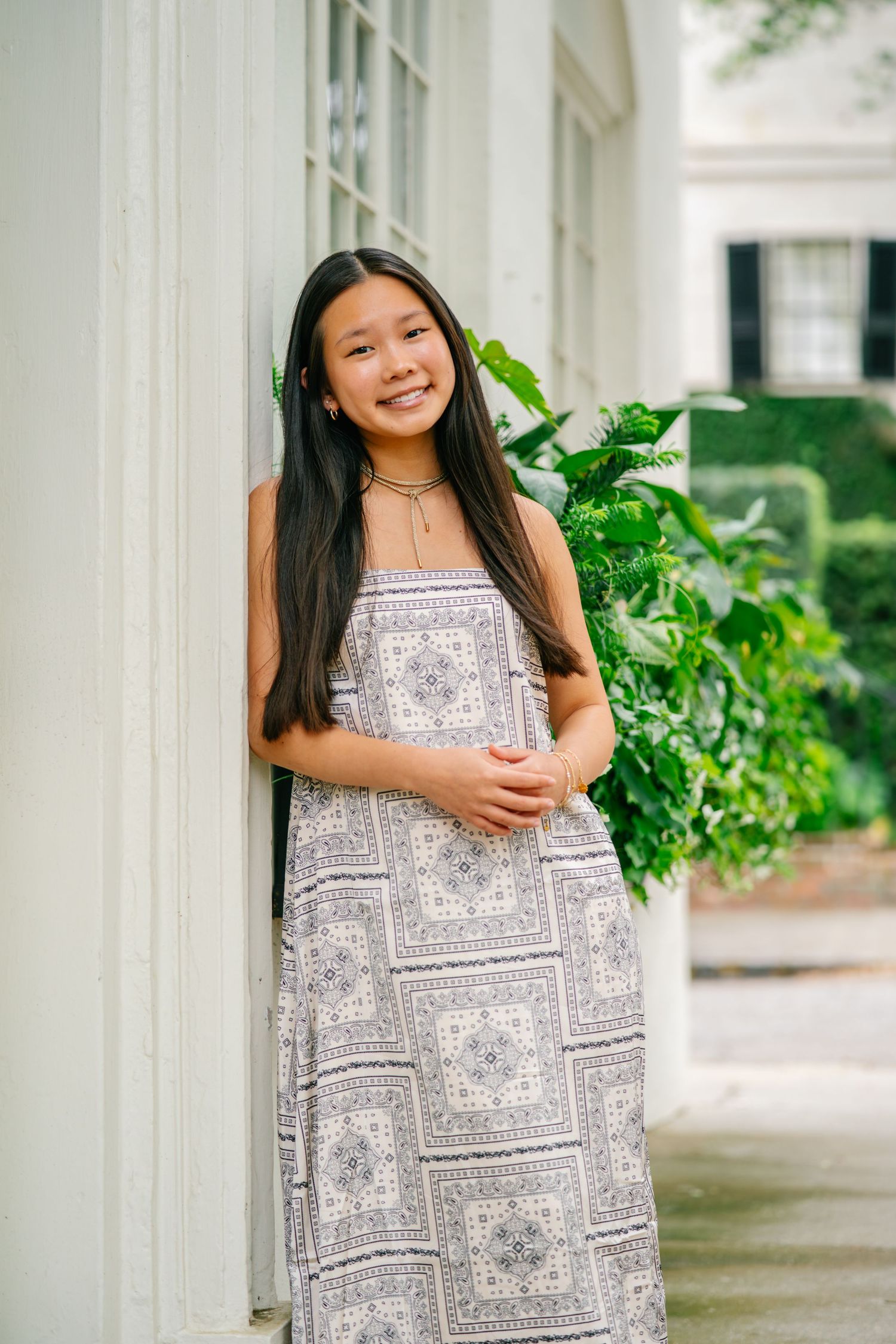 Young girl in patterned maxi dress smiling outdoors near white column and green foliage.