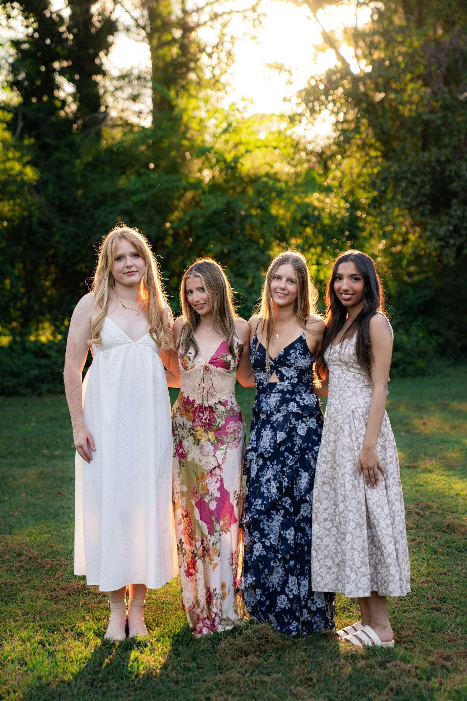 Four young women in summer dresses stand arm in arm on a grassy lawn, smiling in warm golden sunset light with green trees behind.