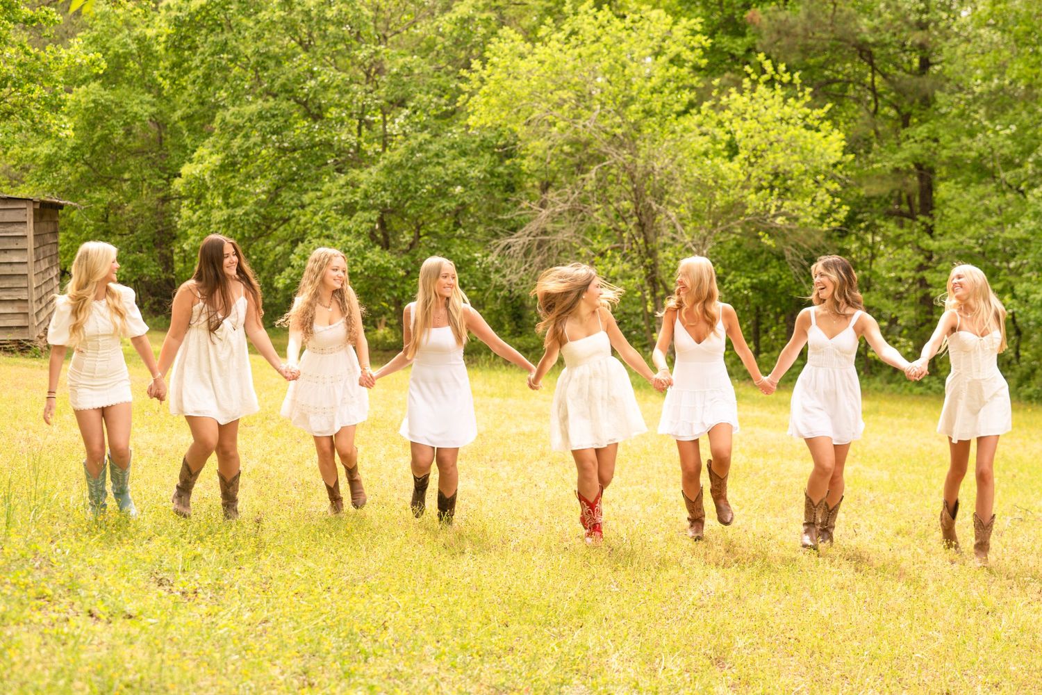 Seven young women in white dresses and cowboy boots hold hands dancing in a sunny green meadow.
