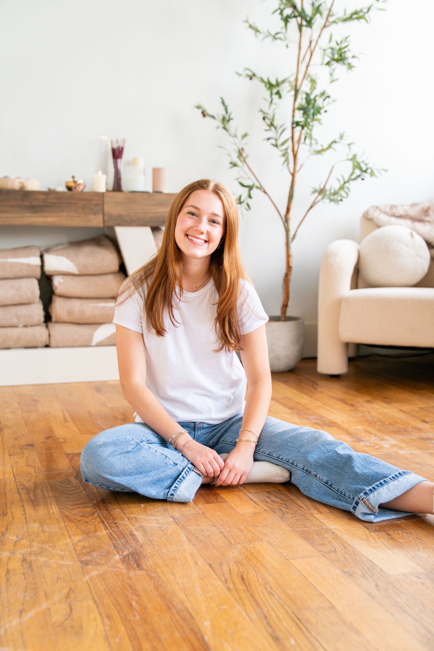 Smiling young woman with long hair sitting cross-legged on hardwood floor in bright living room wearing white tee and jeans.
