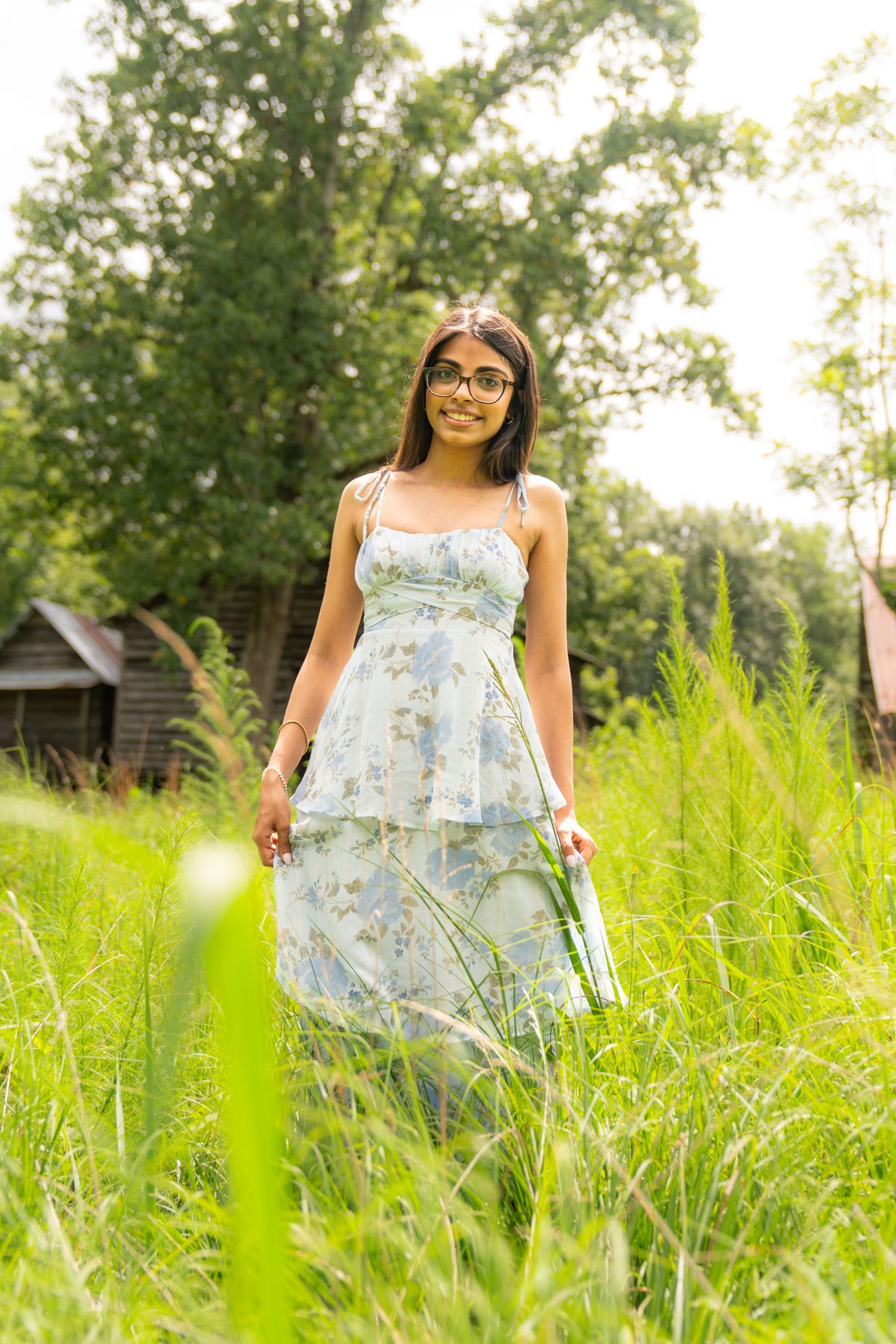 Young woman in floral sundress and glasses standing in tall green grass with trees in background.