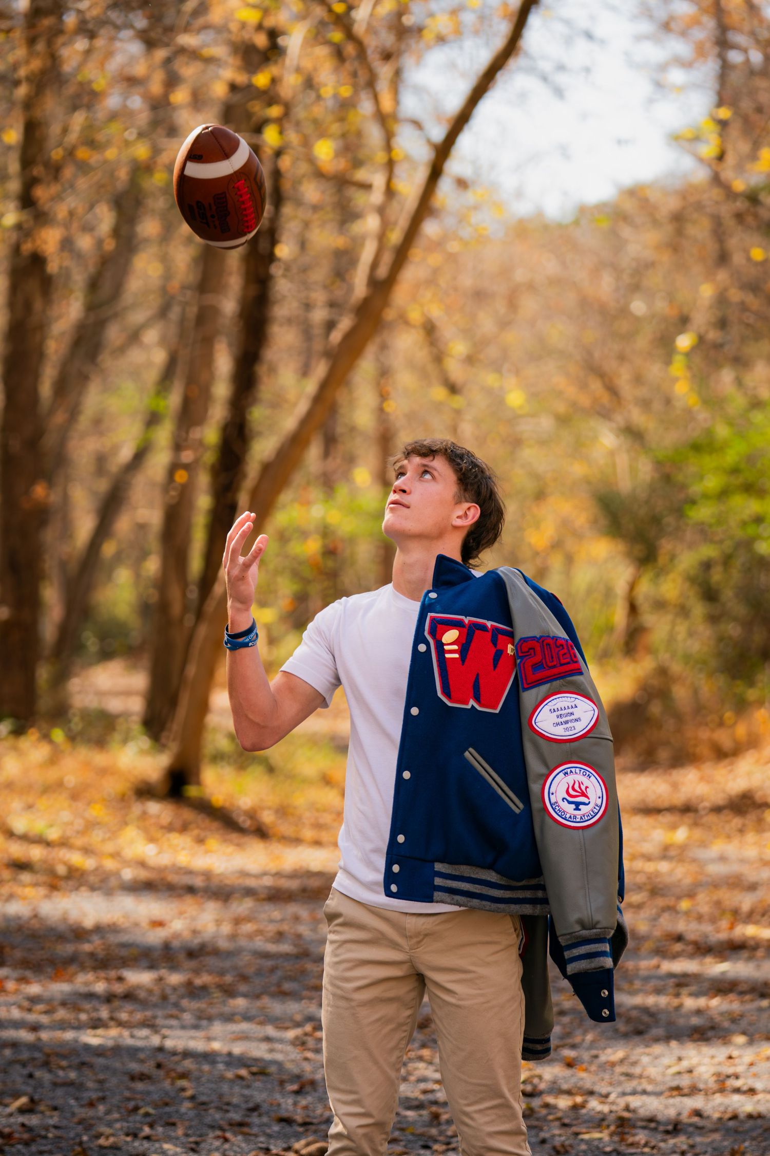 Student tosses football in wooded area while wearing varsity letterman jacket.