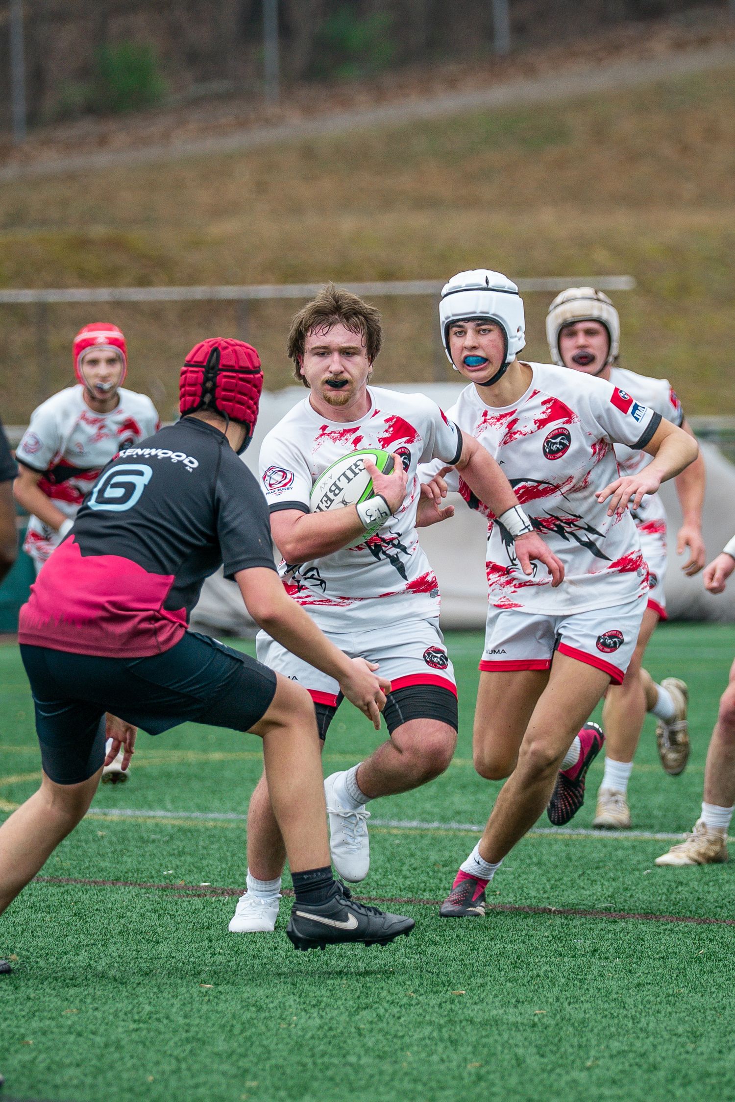 Rugby players compete on artificial turf field with one player carrying ball while opponent attempts defensive tackle.