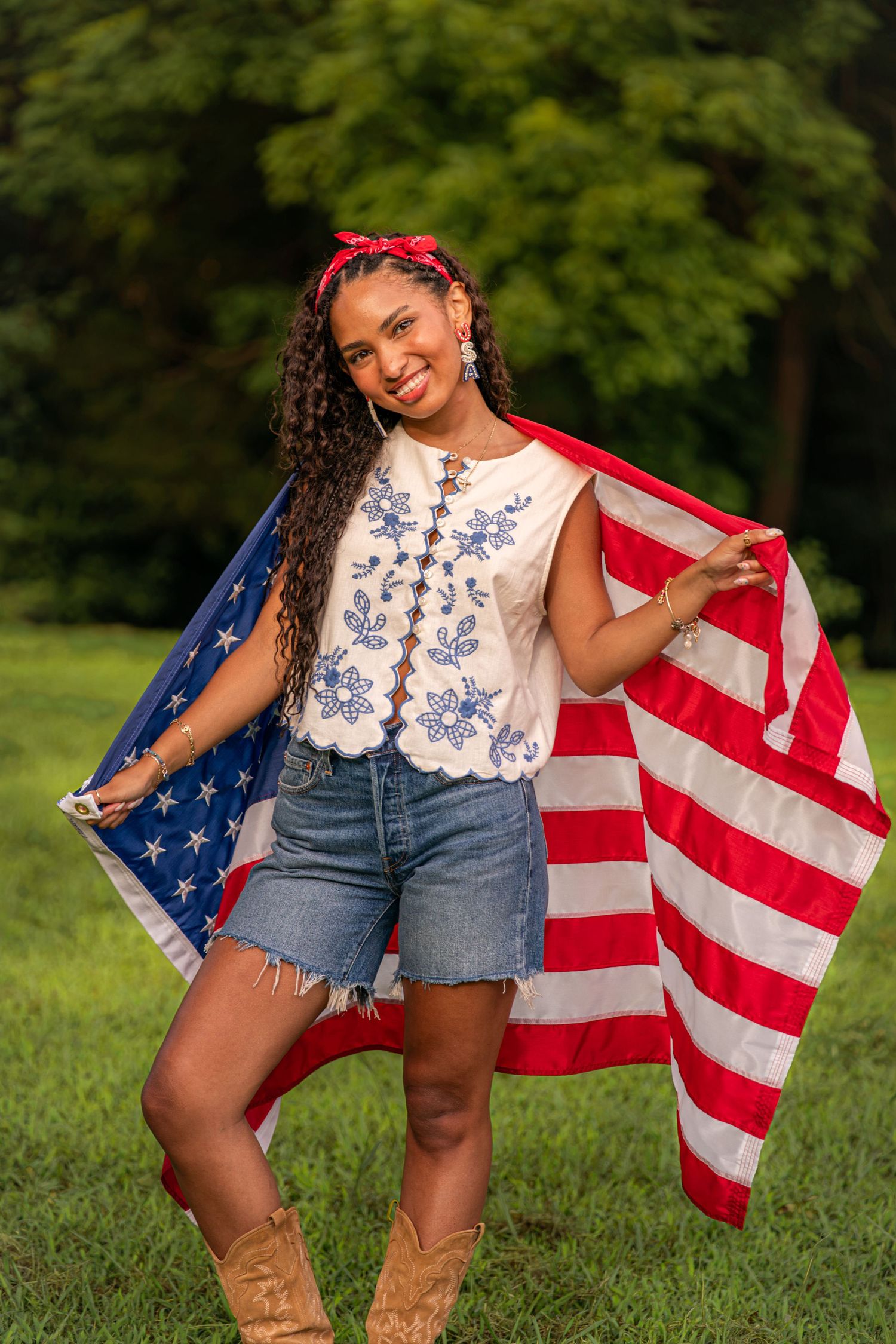 Smiling woman holding American flag outdoors, wearing white floral top, denim shorts, and cowboy boots with red headband.